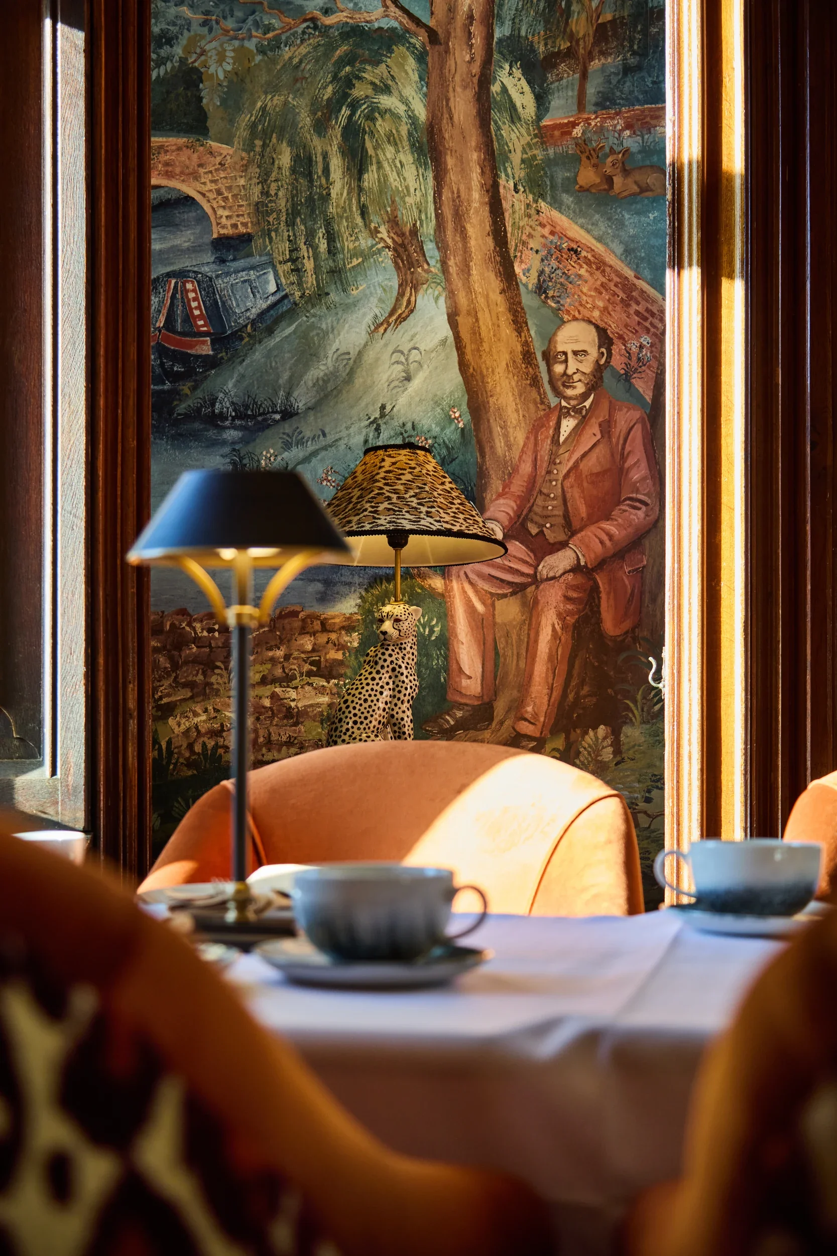 A painting on a wall depicting a pastoral scene with a bridge, trees, a river, and two deer, partially obscured by a table lamp with a leopard print shade, a tiger figurine, and a tea set on a table in the foreground.