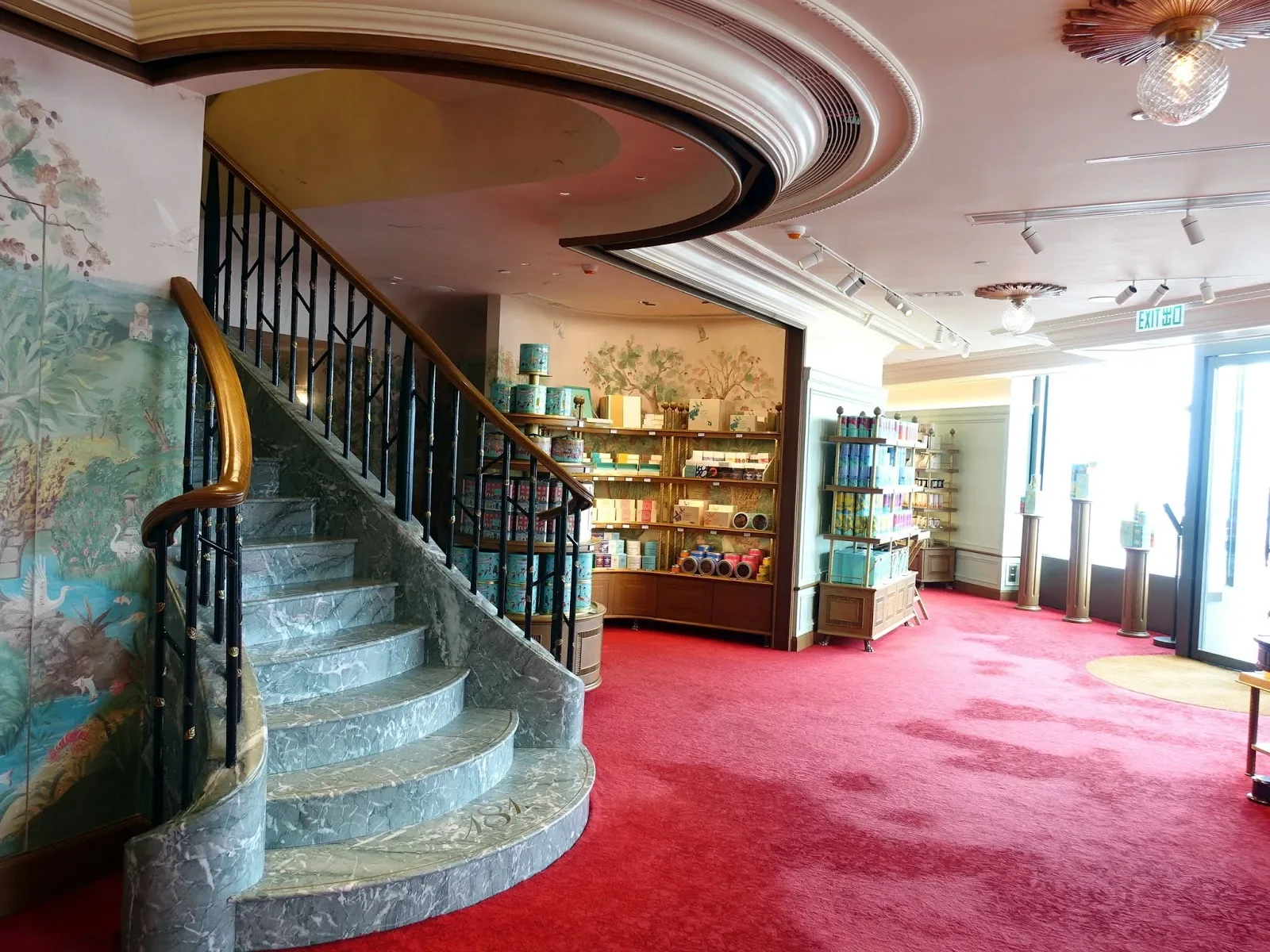 Interior of a building with a curved marble staircase featuring a wooden and black metal railing. The walls behind the stairs are decorated with nature-themed murals. To the right, shelves display colorful products, possibly tissues or decorative boxes, with natural light flooding through large glass doors at the entrance. The ceiling has ornate moldings, light fixtures, and a disco ball, with red carpet flooring.