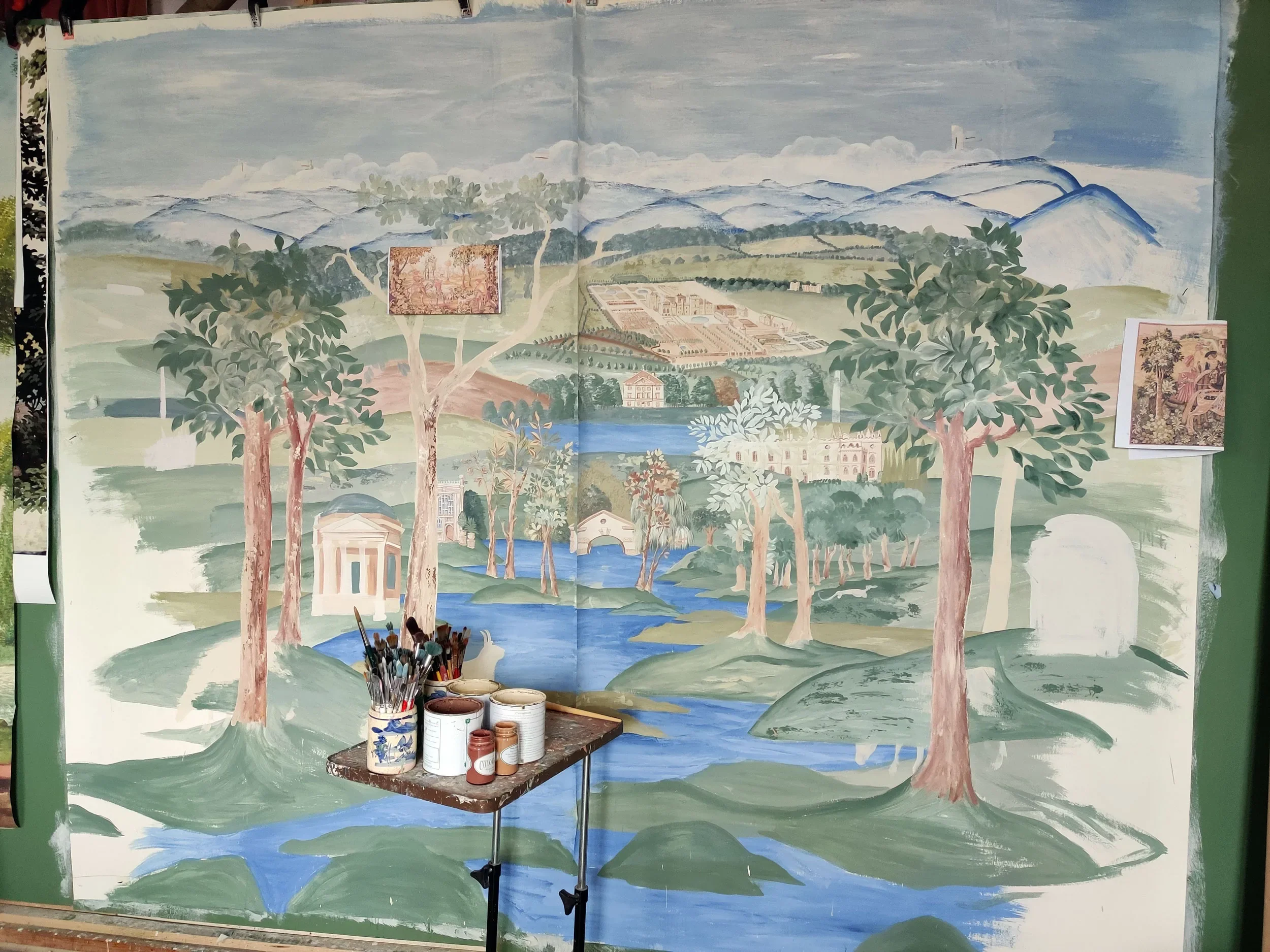 A large mural being painted depicting a landscape with mountains in the background, trees, a river, and classical buildings, with paint cans and brushes in front.