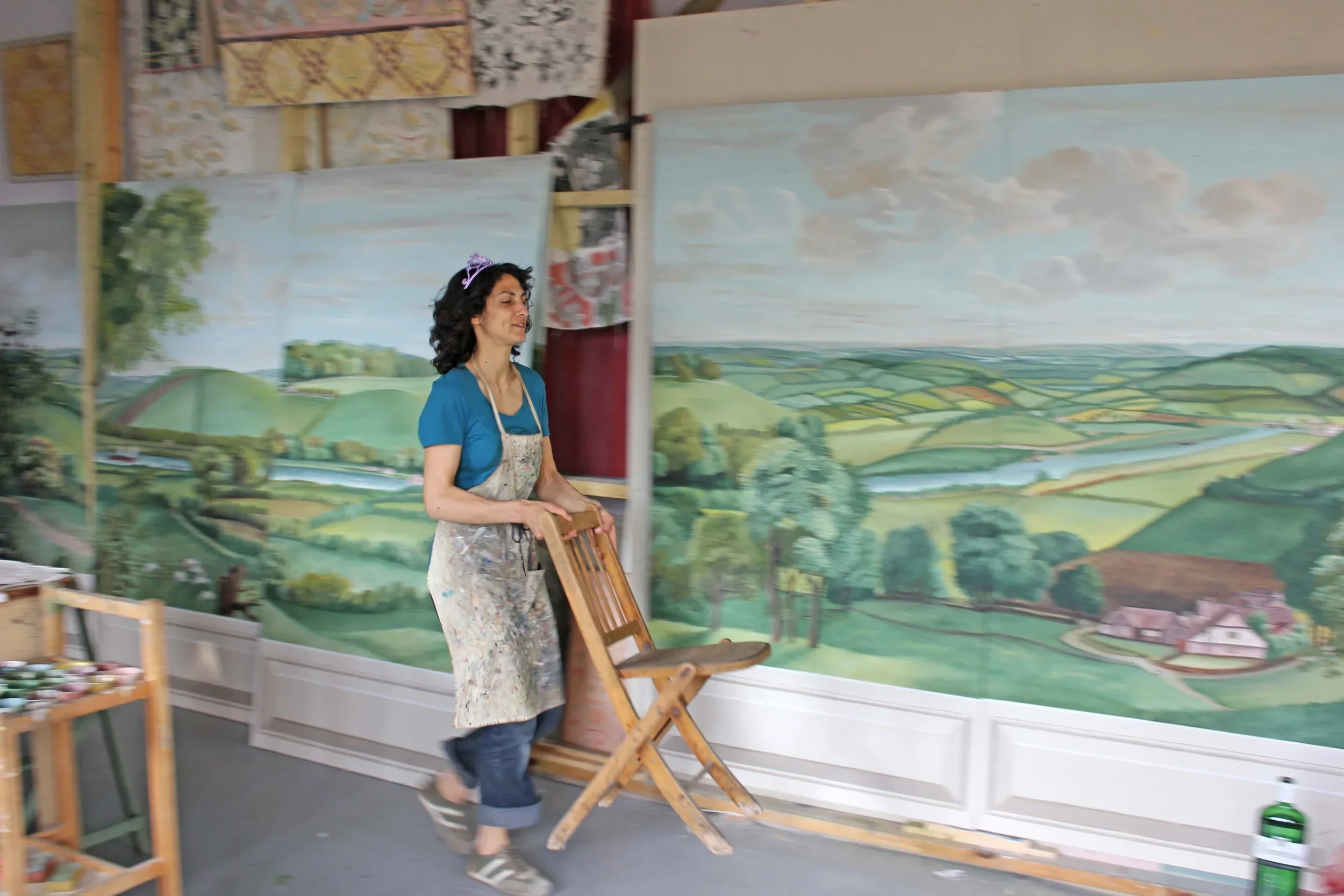 A woman with curly black hair wearing a blue shirt, apron, and clip in her hair, walking barefoot in an art studio, carrying a wooden chair with a landscape mural painted on the background wall.