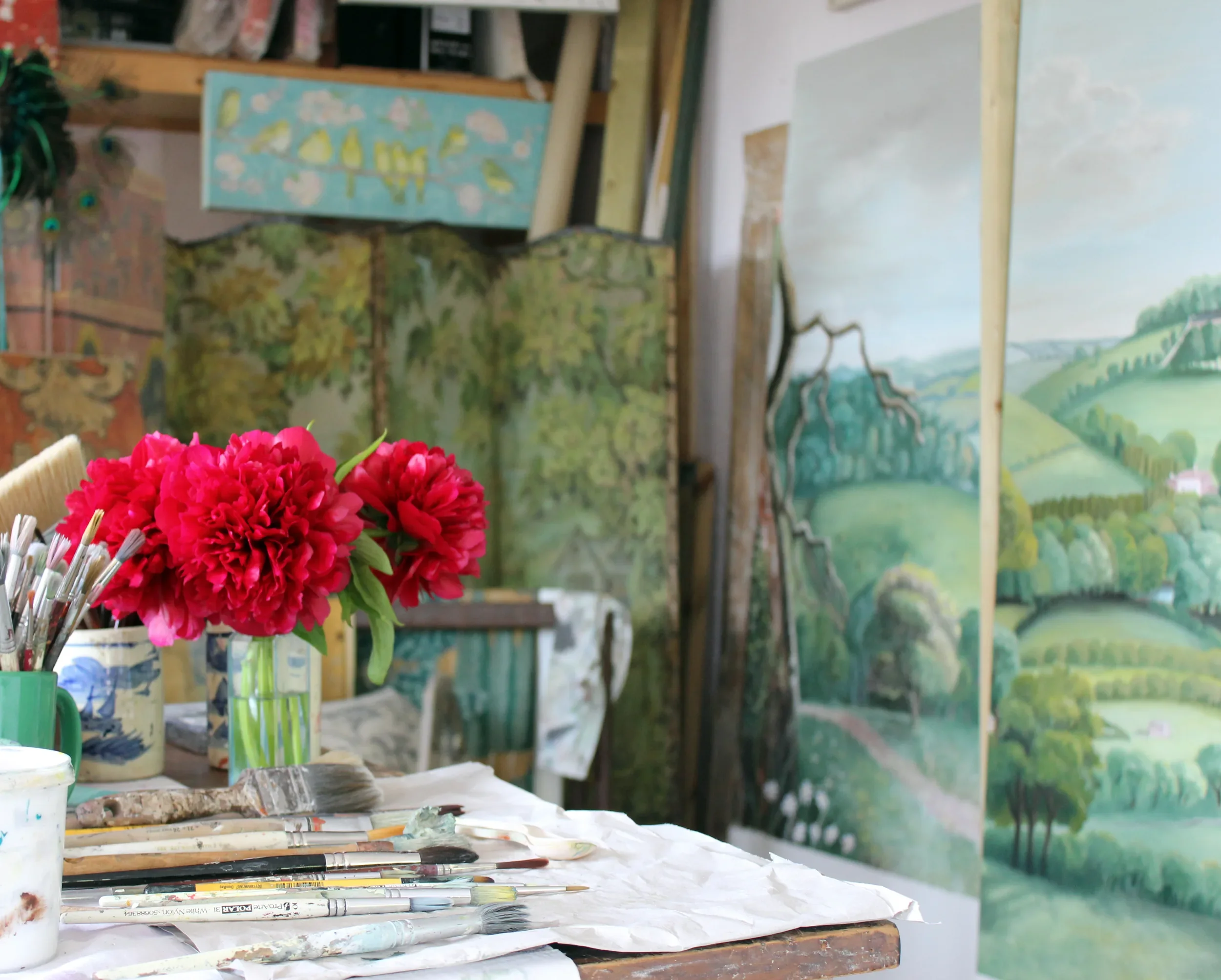 A cluttered art studio workspace with paintbrushes, a glass vase with bright pink flowers, and a background of landscape paintings on the wall.