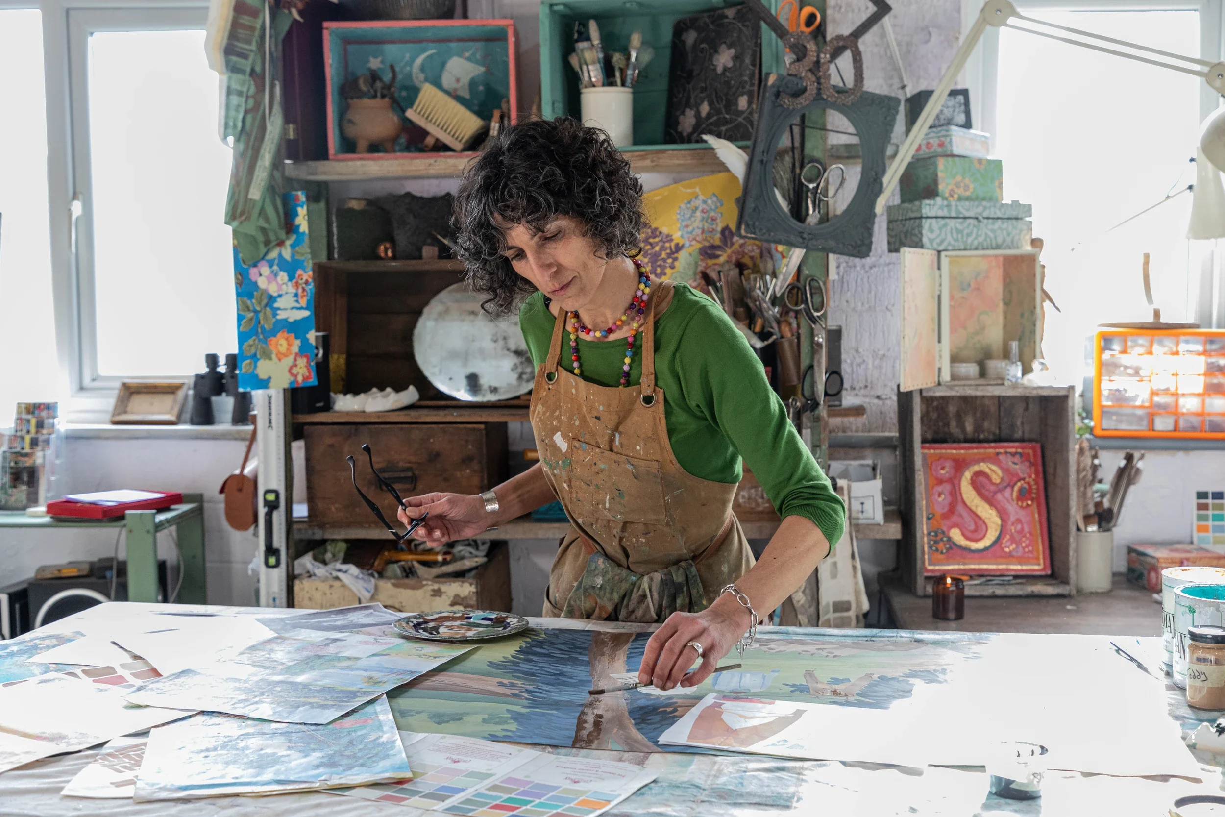Melissa White is pictured working at a large table in her Hastings studio. She is wearing an apron, green jumper and the scene shows a lively studio with many objects including fresco secco.