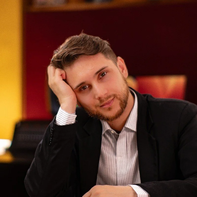 A young man with a beard and brown hair, wearing a black blazer and a striped shirt, resting his head on his hand in a relaxed pose in a warmly lit room.