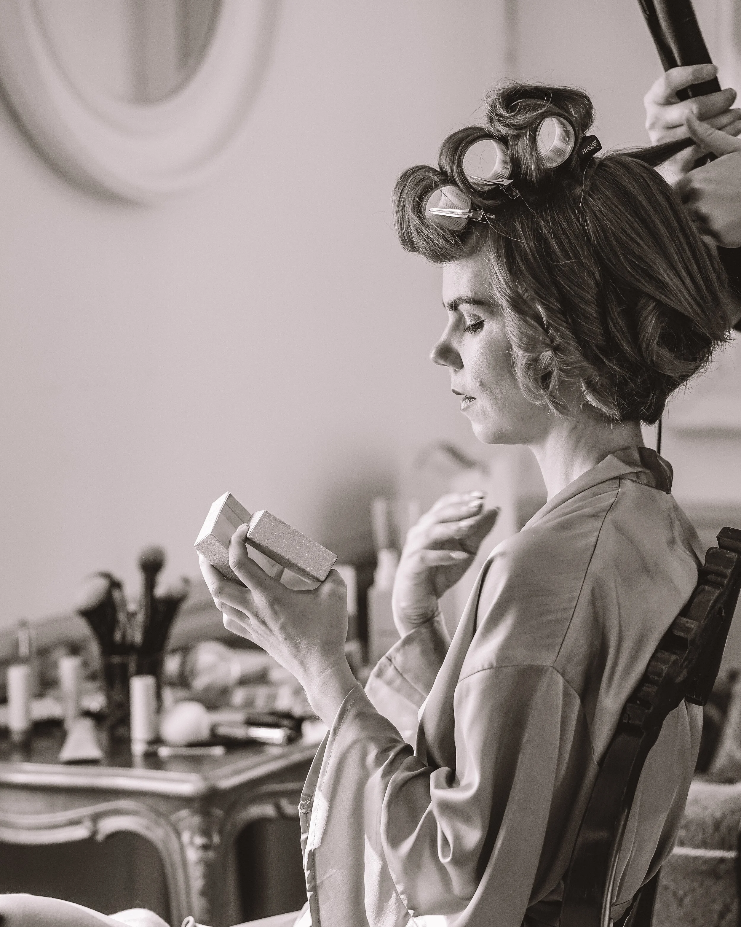 En kvinna med coiffer och hårdlockar sätter sig i en frisörstol, ser på en liten spegel, och blir förberedd för hårstyling.