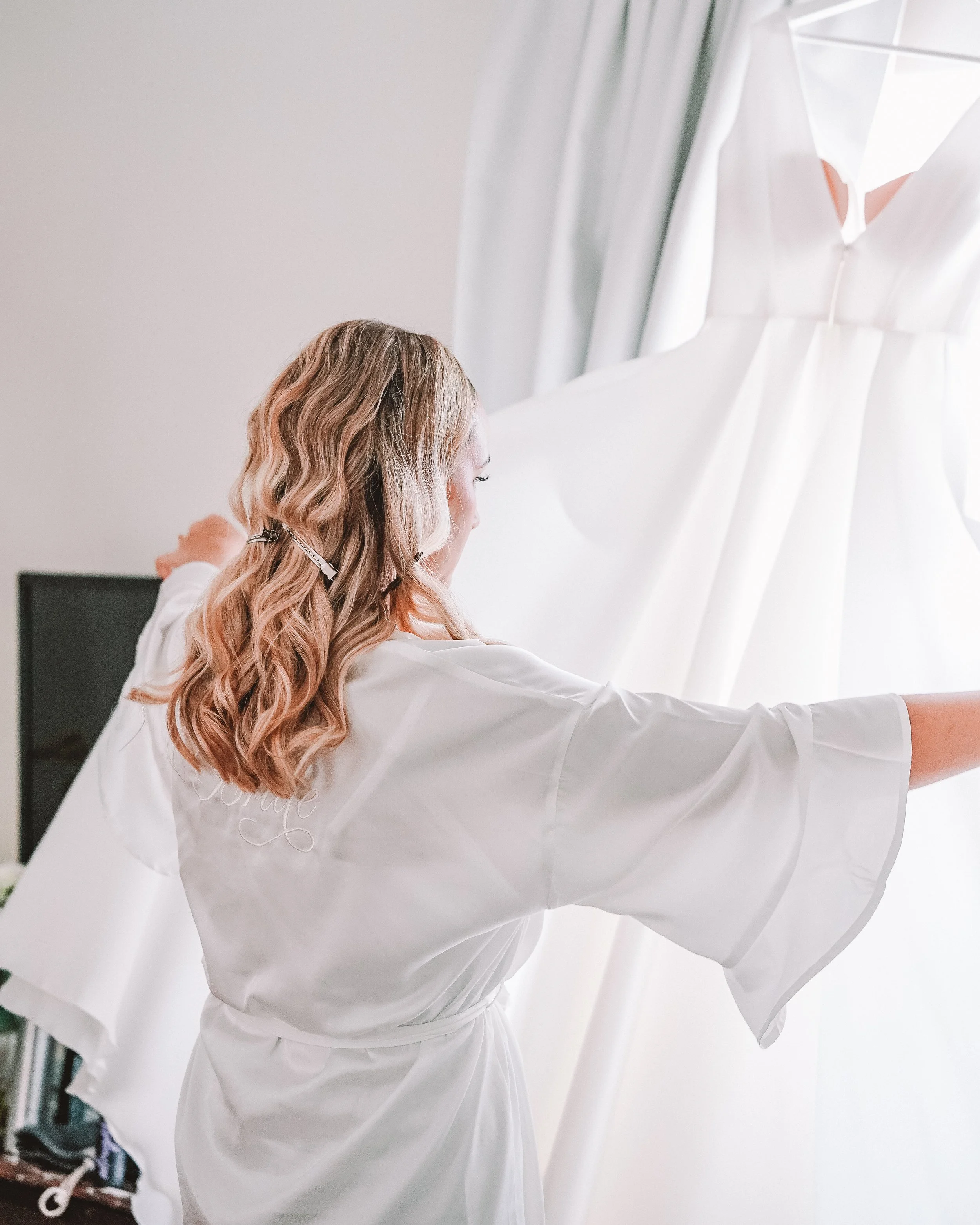 En kvinna i ett vitt satinlakan provar en brudklänning foran en spegel.