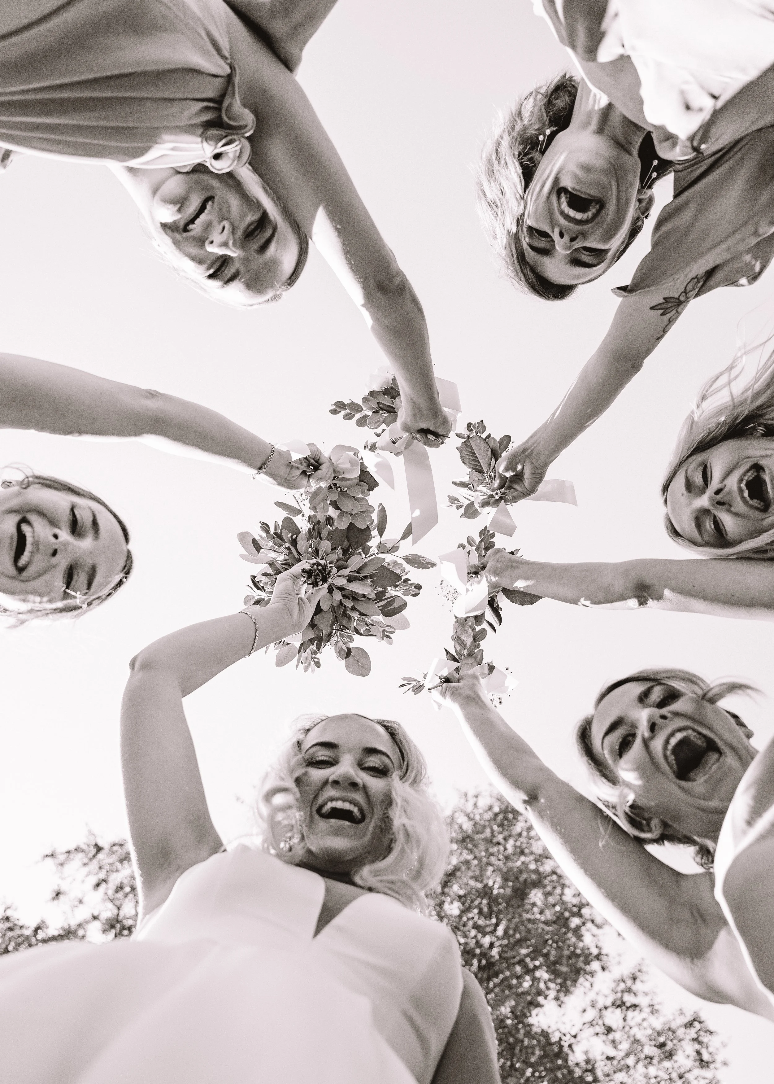 En grupp kvinnor som skrattar och håller i blomsterarrangemang mot en himmel.