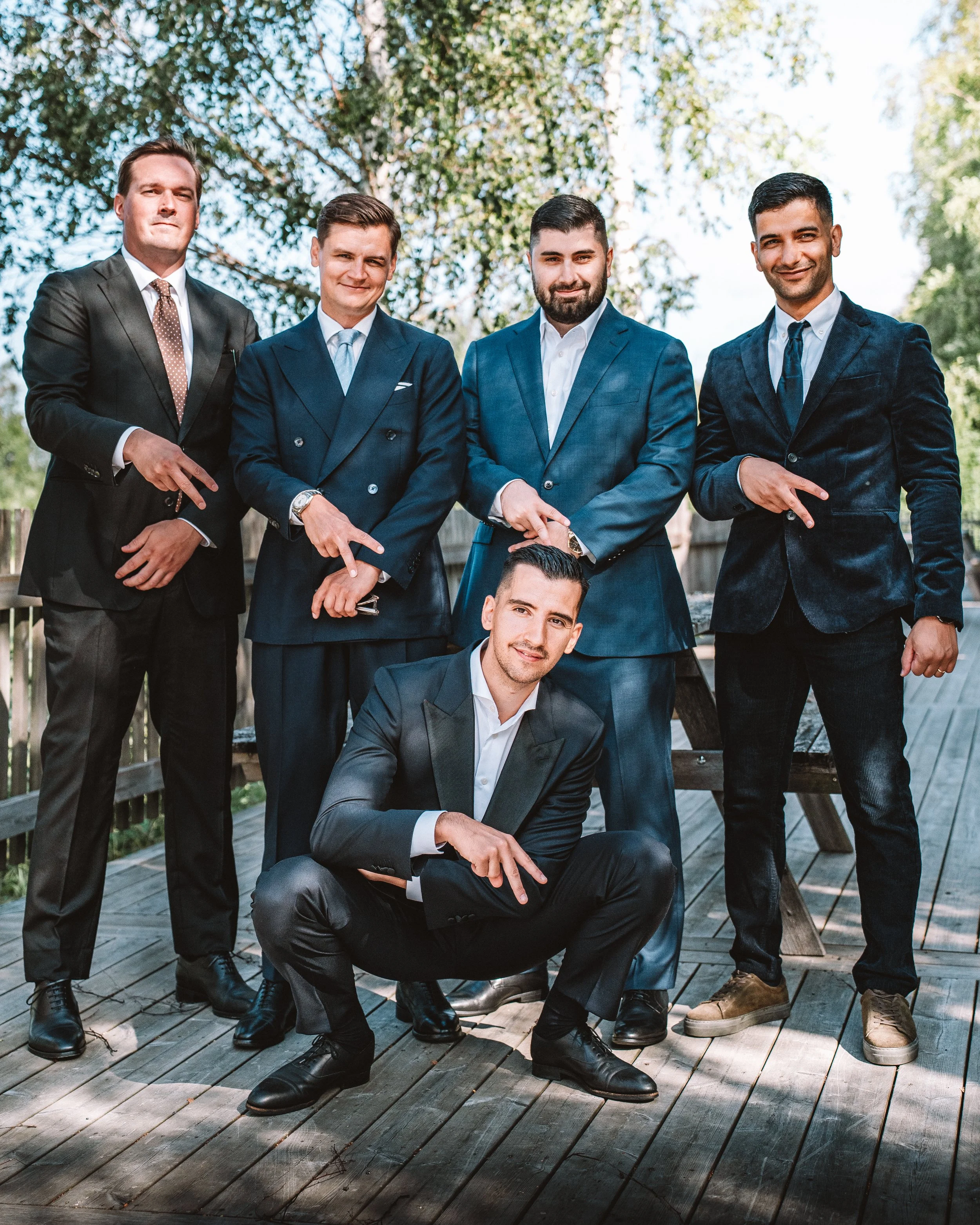 Grupp av fem män i kostymer utomhus, fotograferade i linje och en knästående, alla gör peace-tecken med händerna på en träborg.