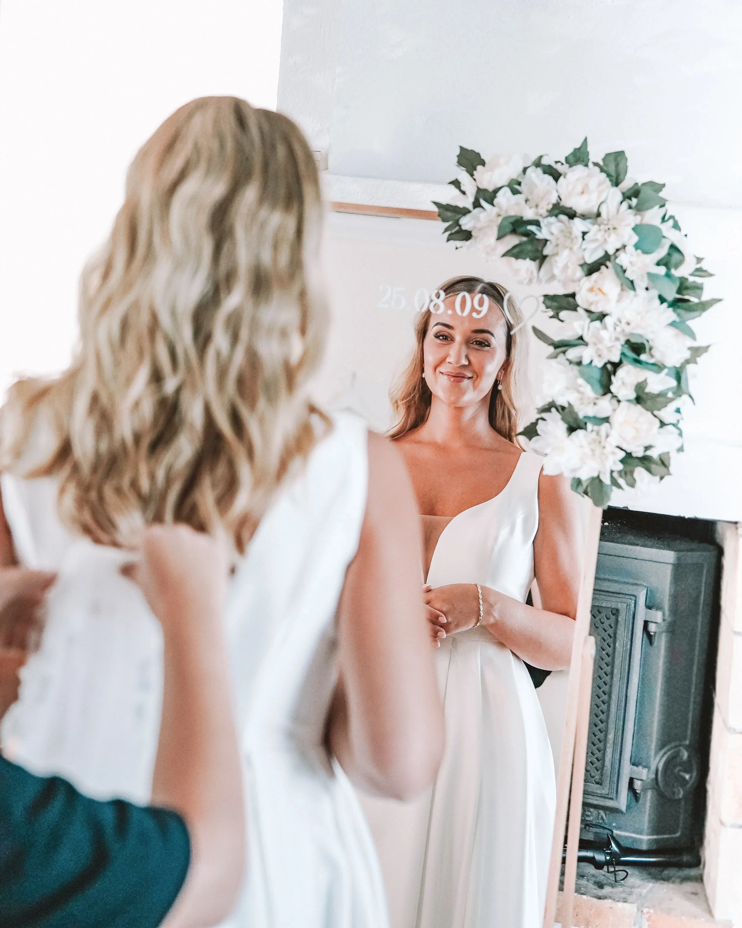 En brud i vit brudklänning ser sig själv i en spegel framför en blomsterdekor.