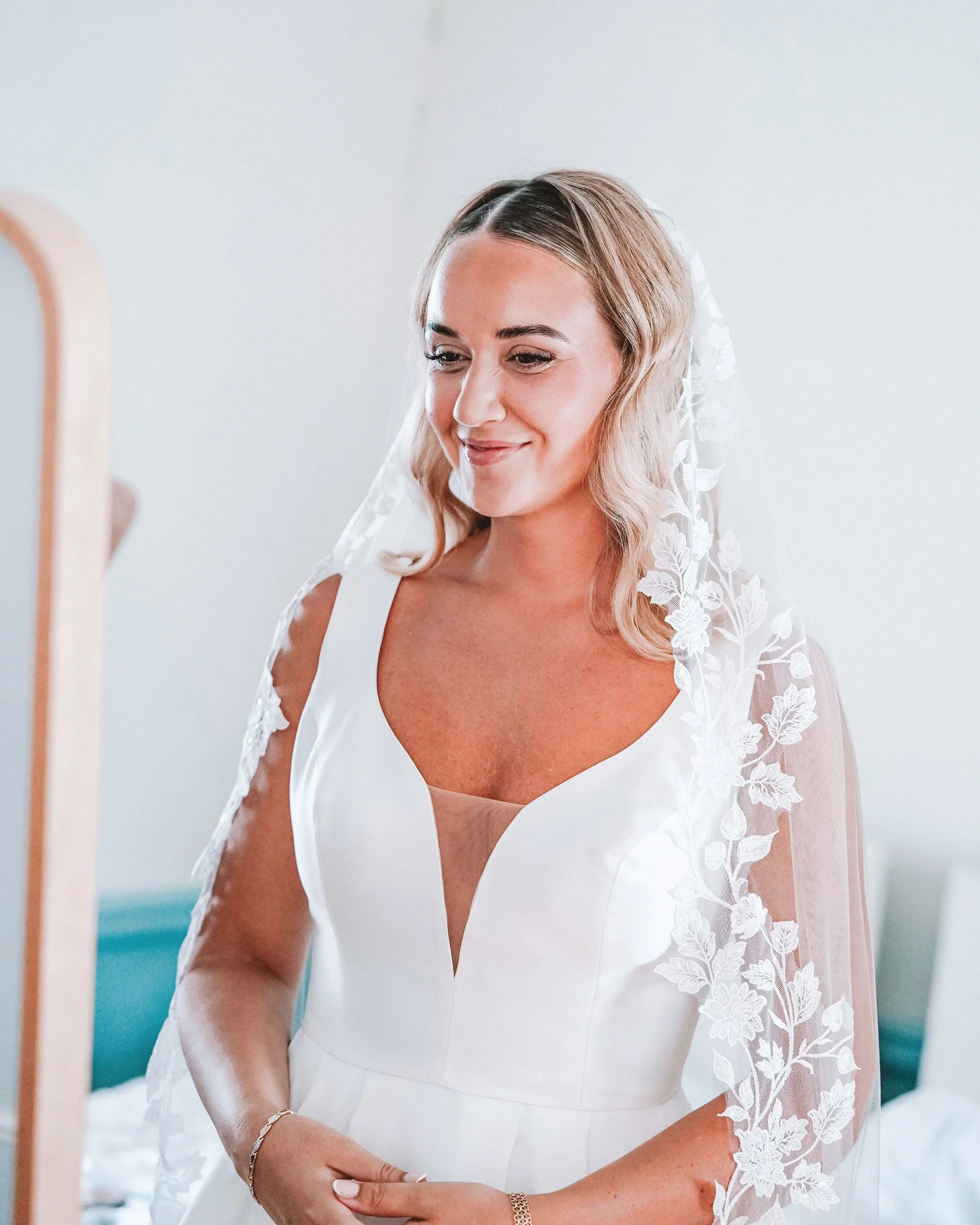 En kvinna på ett bröllop, klädd i en vit brudklänning med spetsdetaljer och slöja, ser i spegel.
