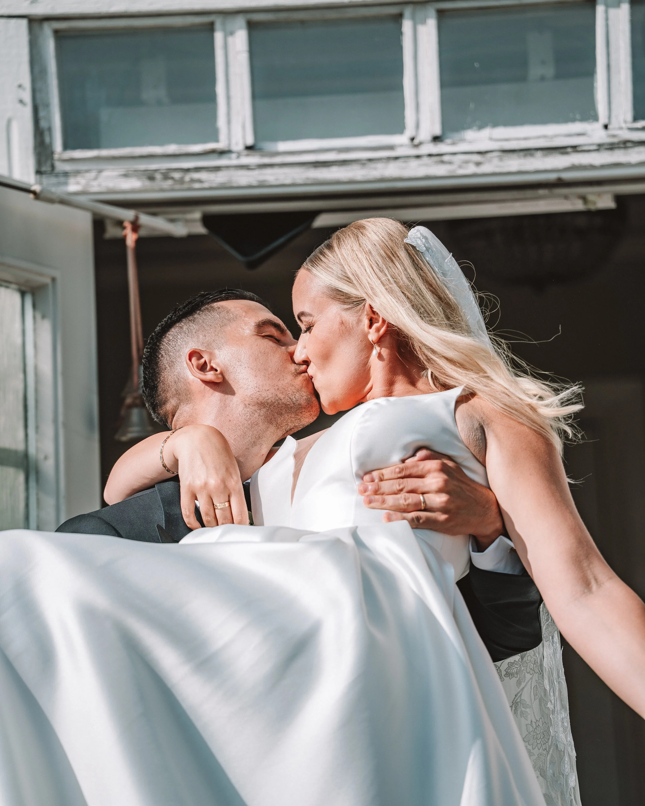 En nygiften man och kvinna kysser varandra under deras bröllop, mannen bär en mörk kostym och kvinnan en vit brudklänning med slöja.