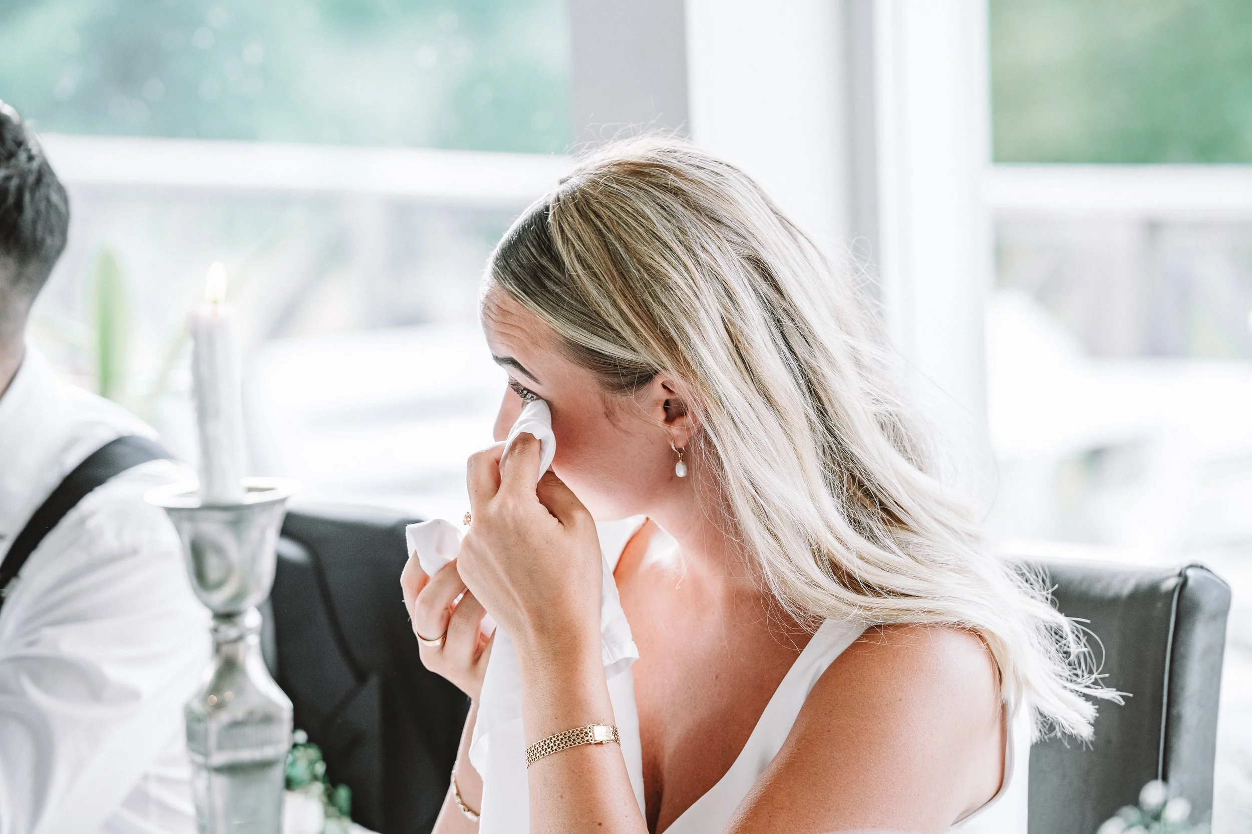 En kvinna med blont hår torkar tårar med en näsduk vid ett bord inomhus, klädd i vit klänning.