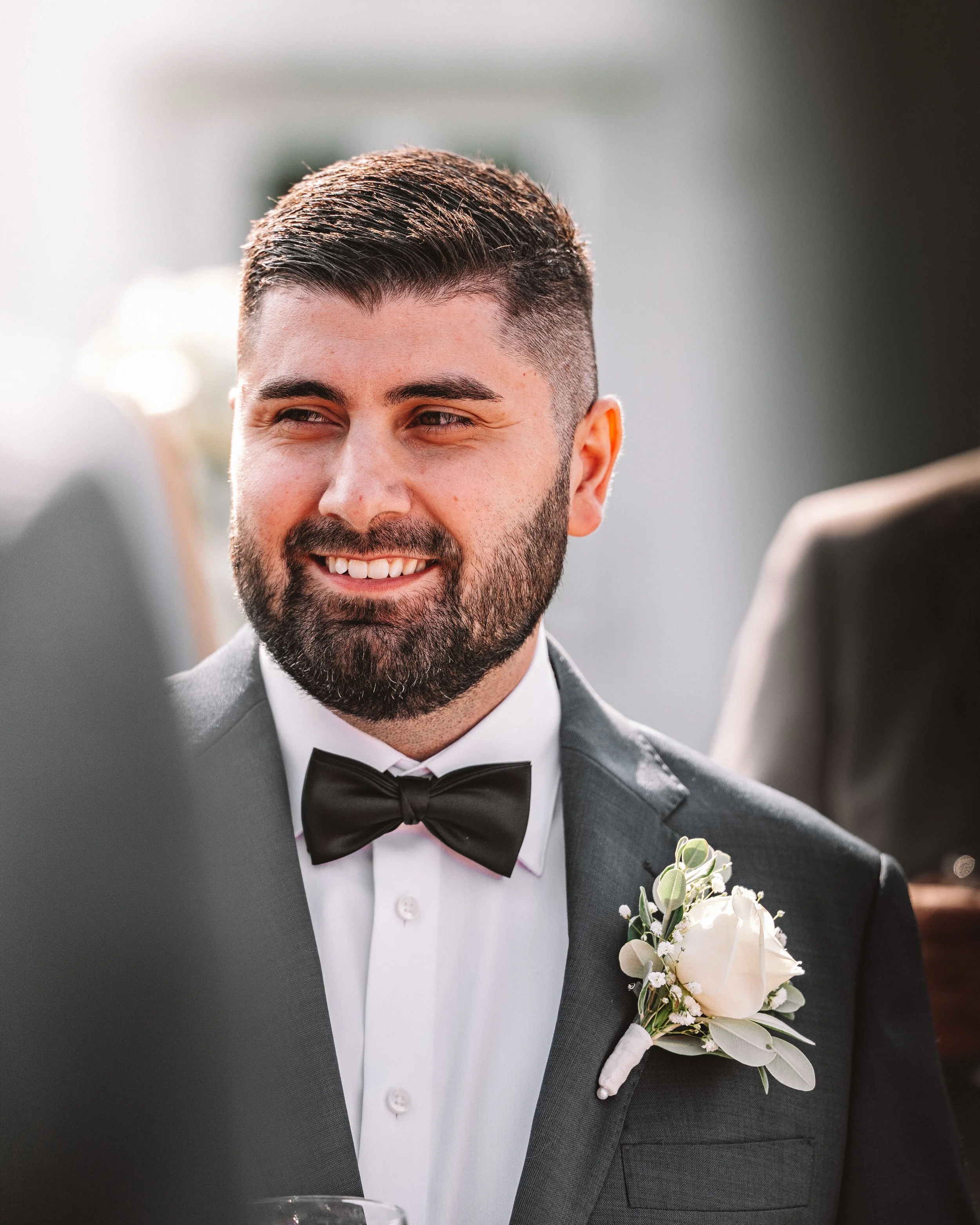 En man i svart tuxedo och svart rosett med vit skjorta, leende, med en vit blomsterboutonnière på kavajén, under ett formellt tillfälle.