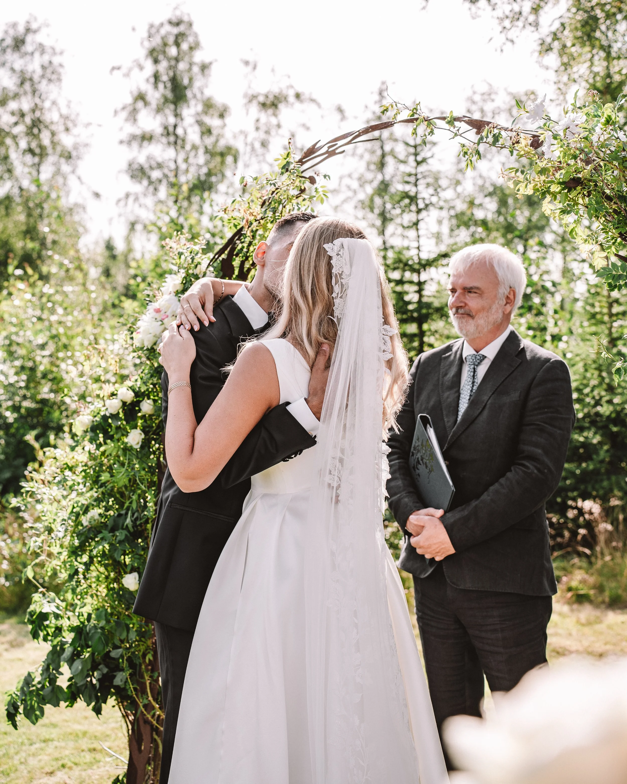 Ett brudpar kysser varandra under ett vigselarrangemang, med en trolig vigselförrättare som håller ett anteckningsblock.