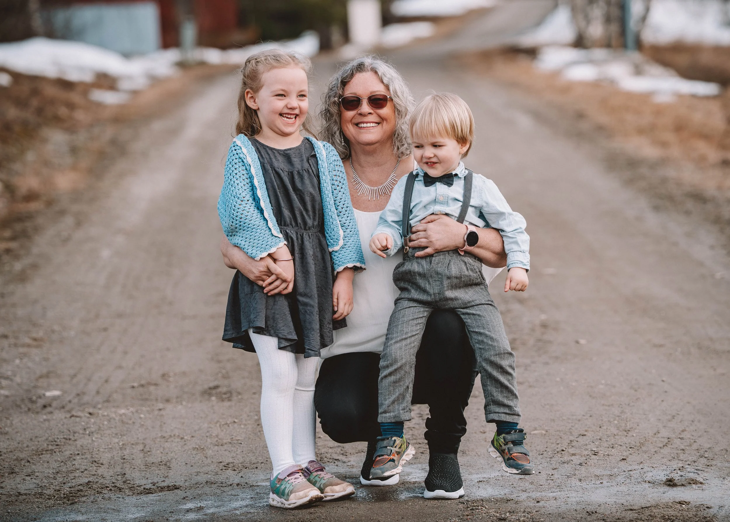 En kvinna med två barn, ett flickebarn och ett pojkbarn, står på en landsväg med vinterlandskap i bakgrunden I Örnsköldsvik, Höga Kusten. Alla ler och ser glada ut.