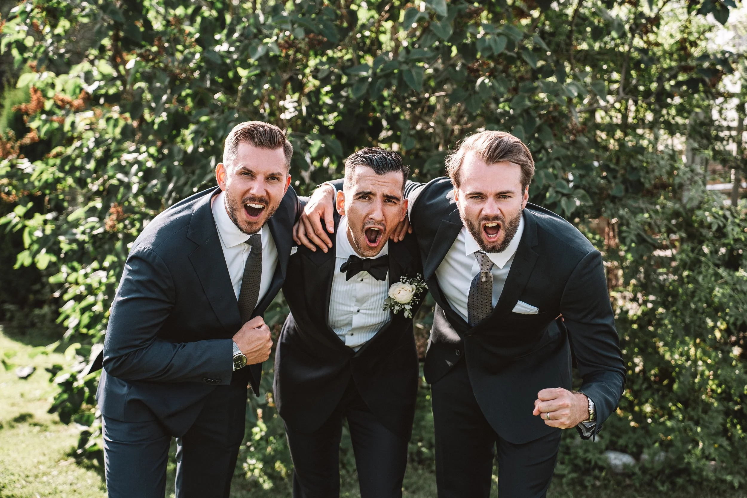 Tre män i kostymer utomhus som skriker och poserar energiskt.