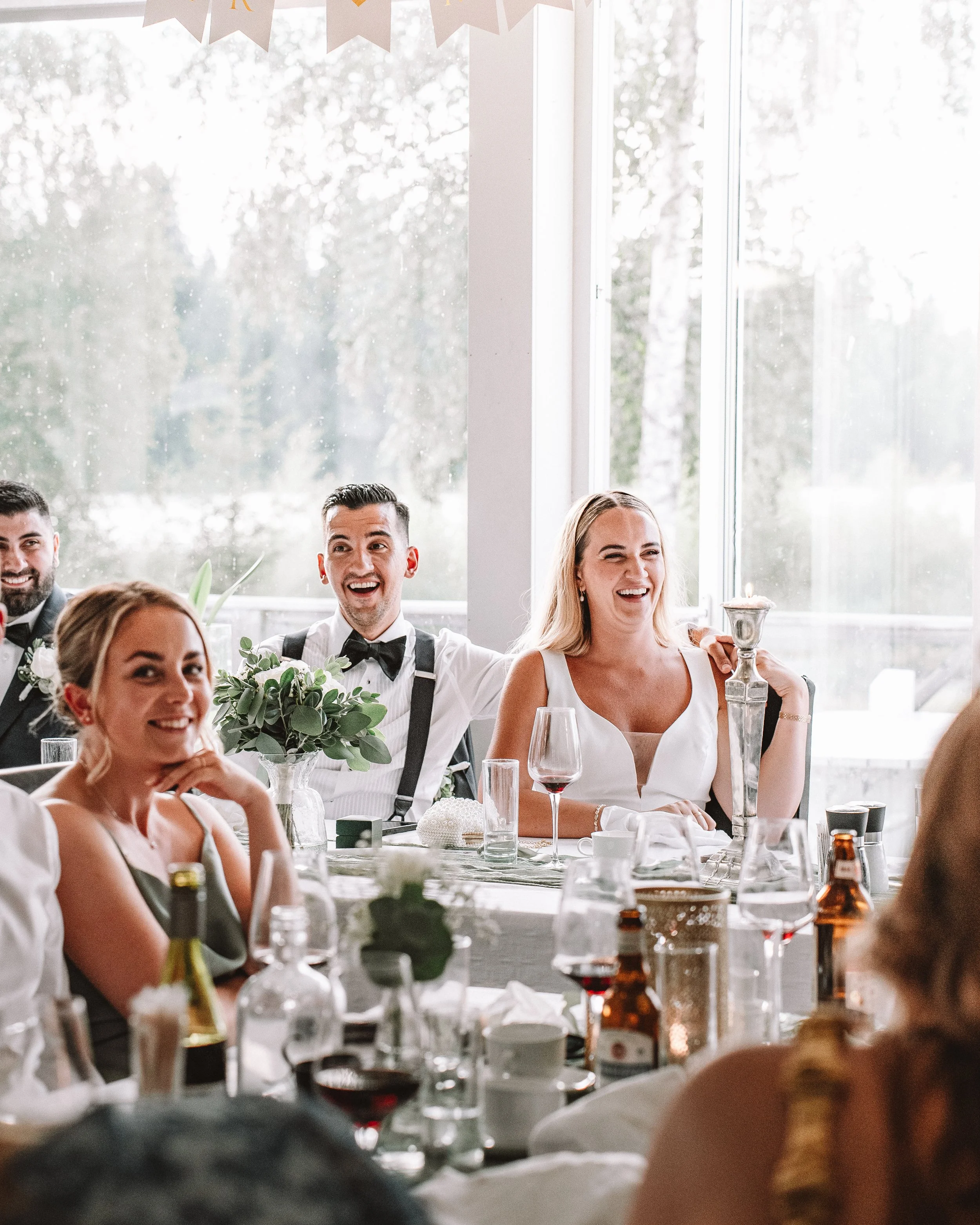 Glada människor sitter vid festbord, troligen på ett bröllop, med blommor och drinkar.