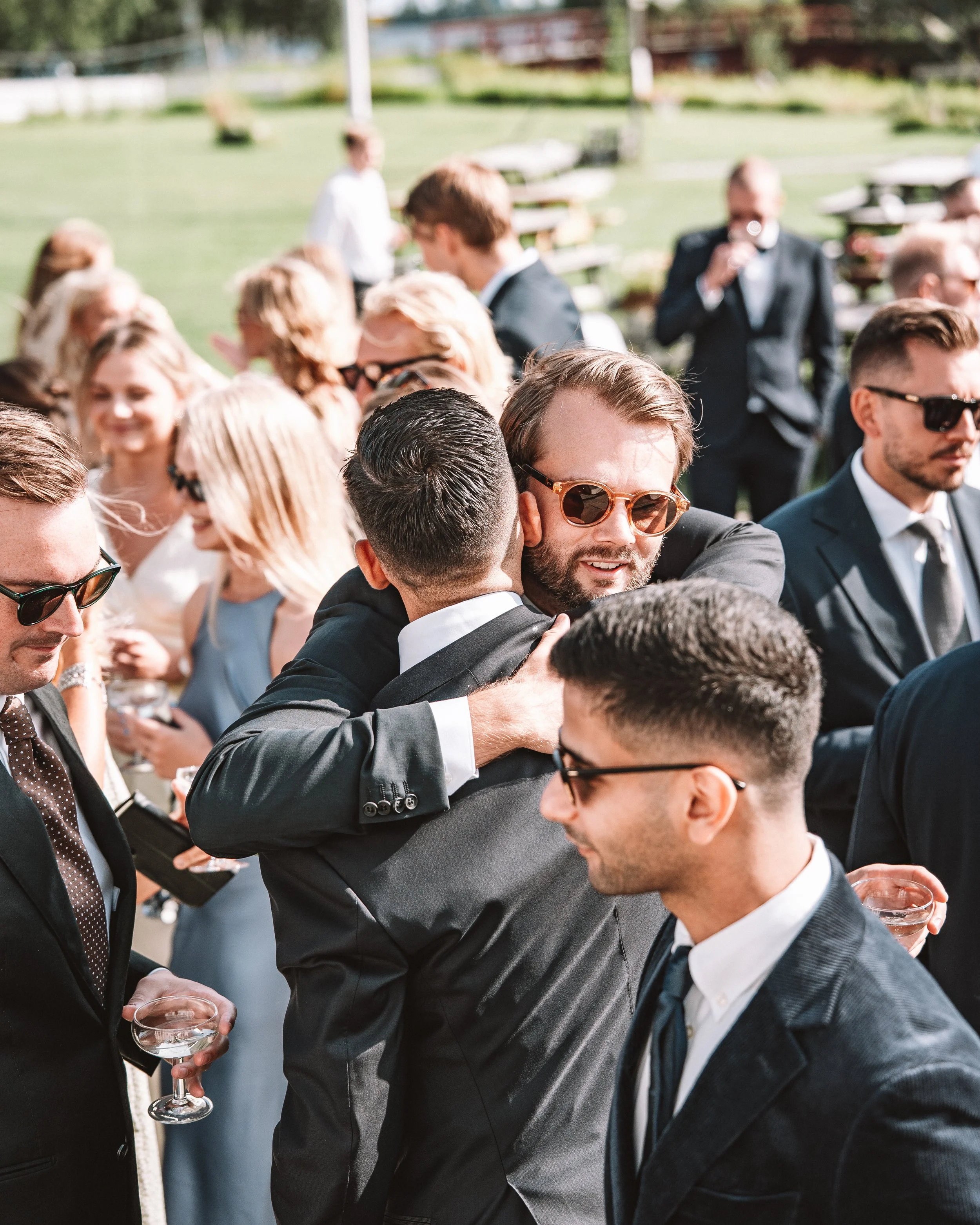 Människor som engagerat kramas och pratar på ett utomhusarrangemang, många bär formella kläder och solglasögon