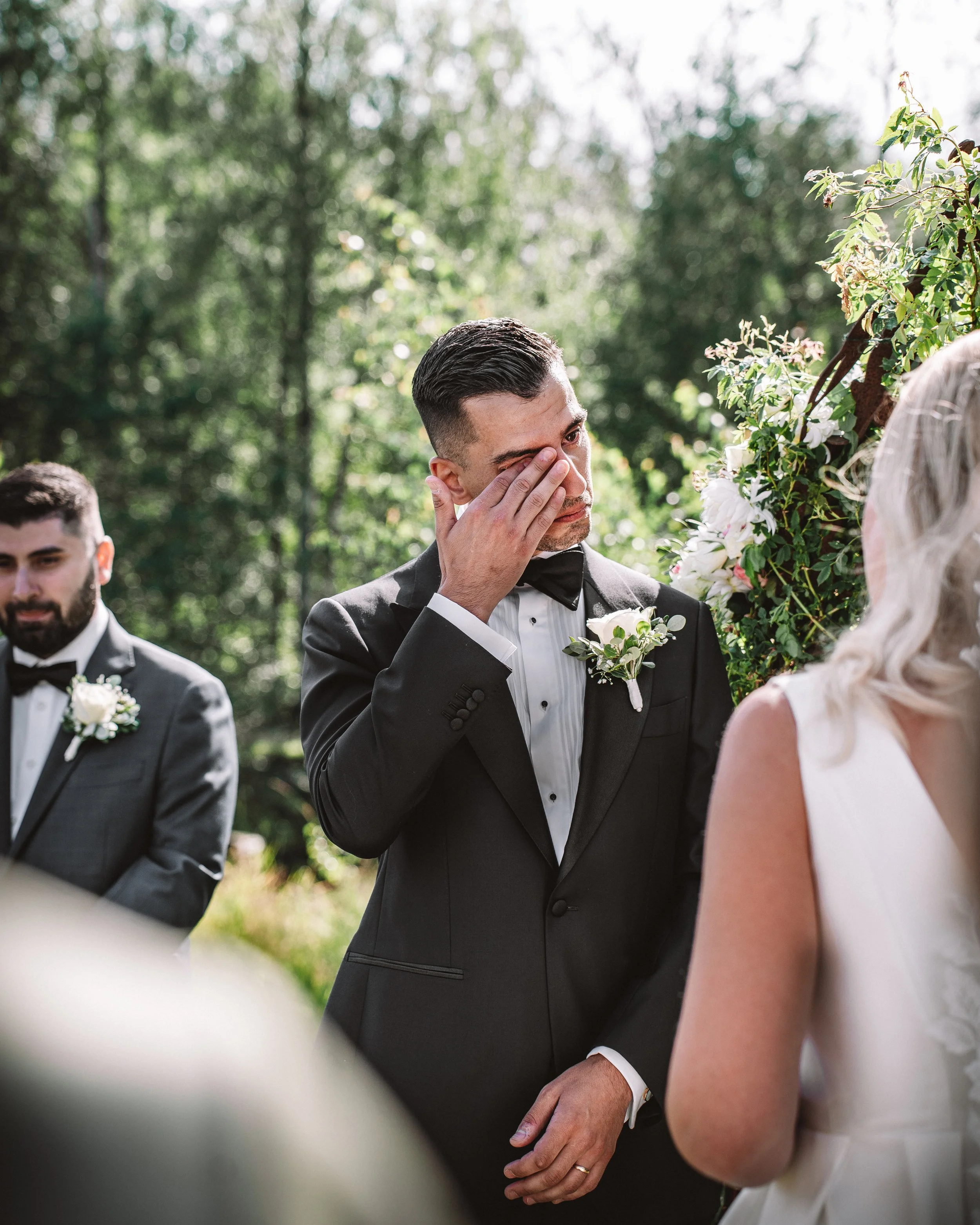 En man i smoking, som verkar gråta, står vid ett bröllop utomhus med träd i bakgrunden. Han är troligtvis brudgummen, och andra personer i formella kläder är också närvarande.