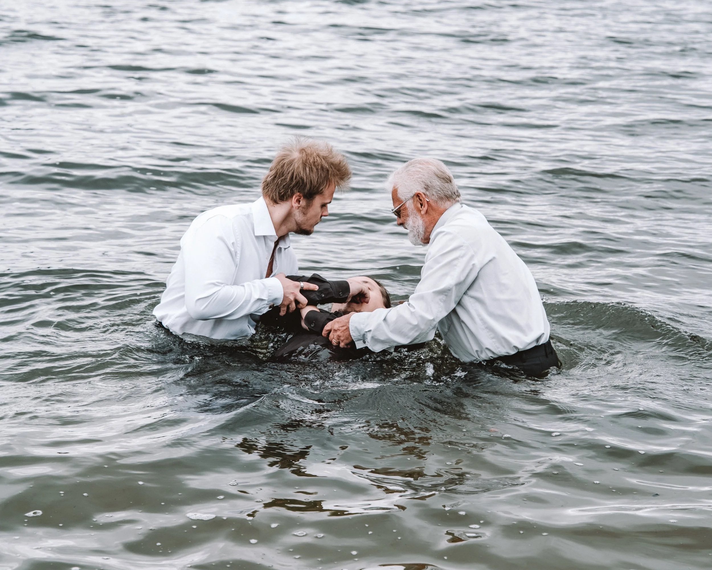 Två män sänker ner en ung man i vattnet, som låter sig döpas i en sjö eller hav, de använder vita skjortor, och den äldre mannen har grått hår och skägg.