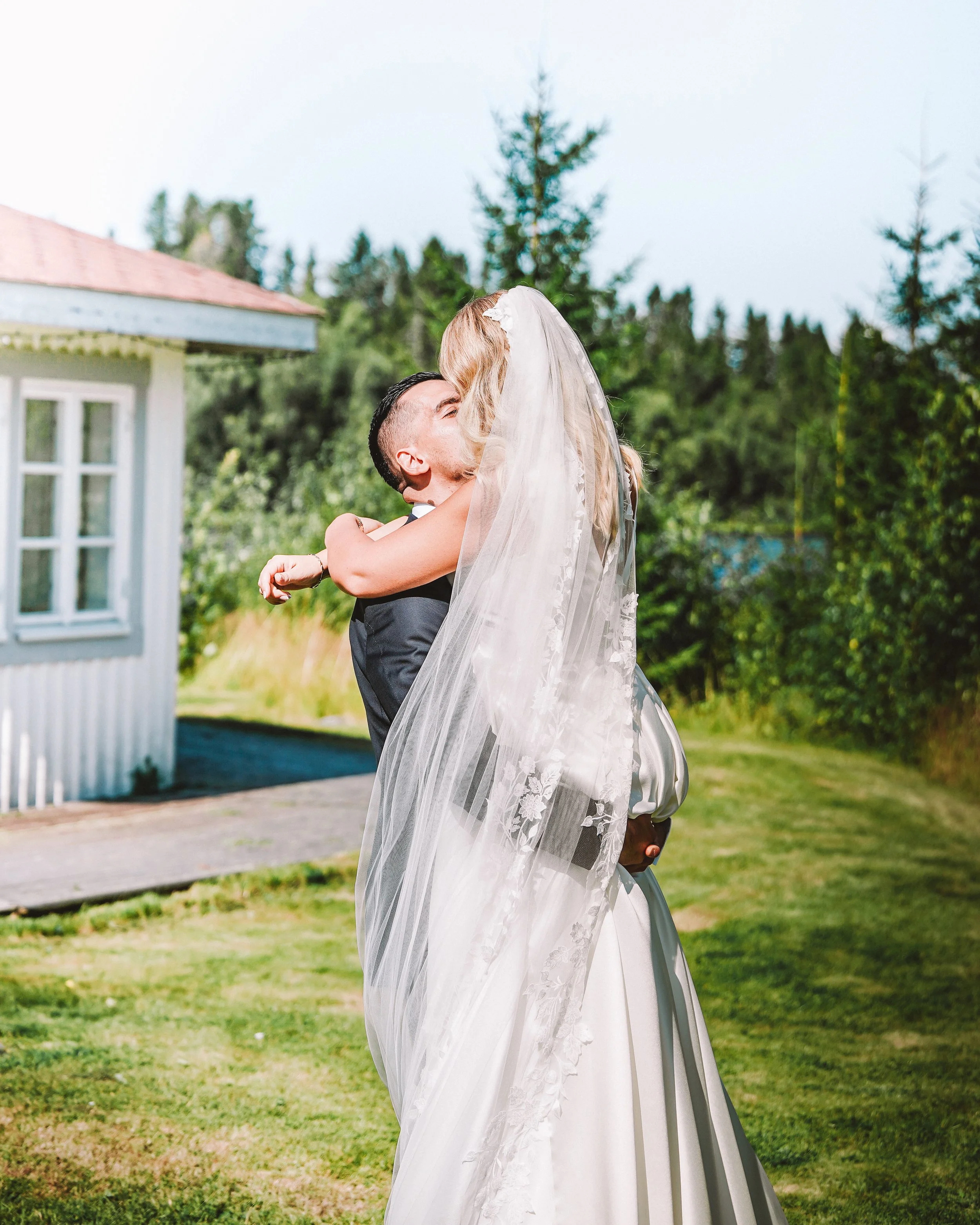 En brud och en brudgums i en kyss utomhus under en ceremonin, med en vit byggnad och gröna träd i bakgrunden.