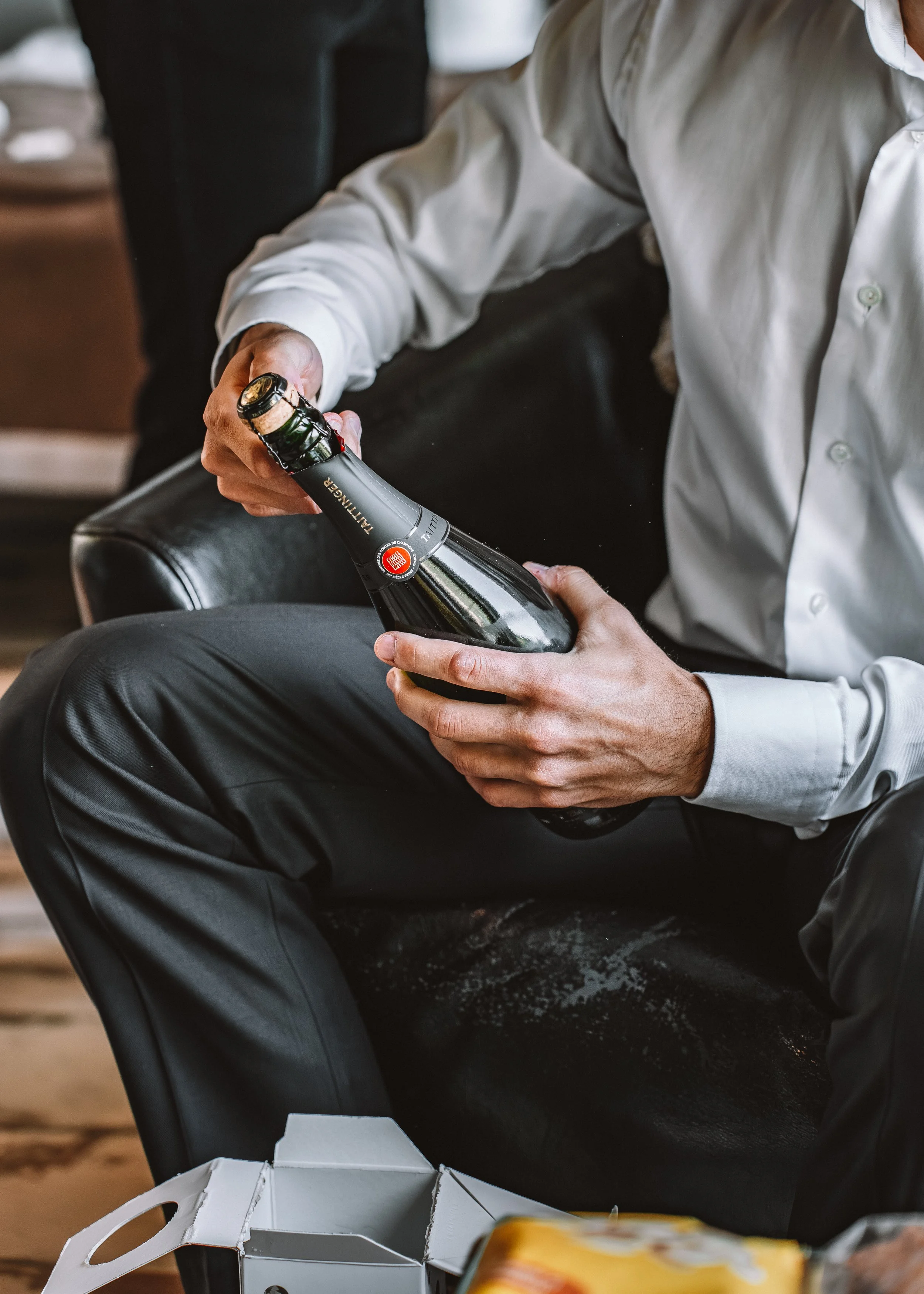 En man i vit skjorta sitter i en soffa och öppnar en flaska champagne.