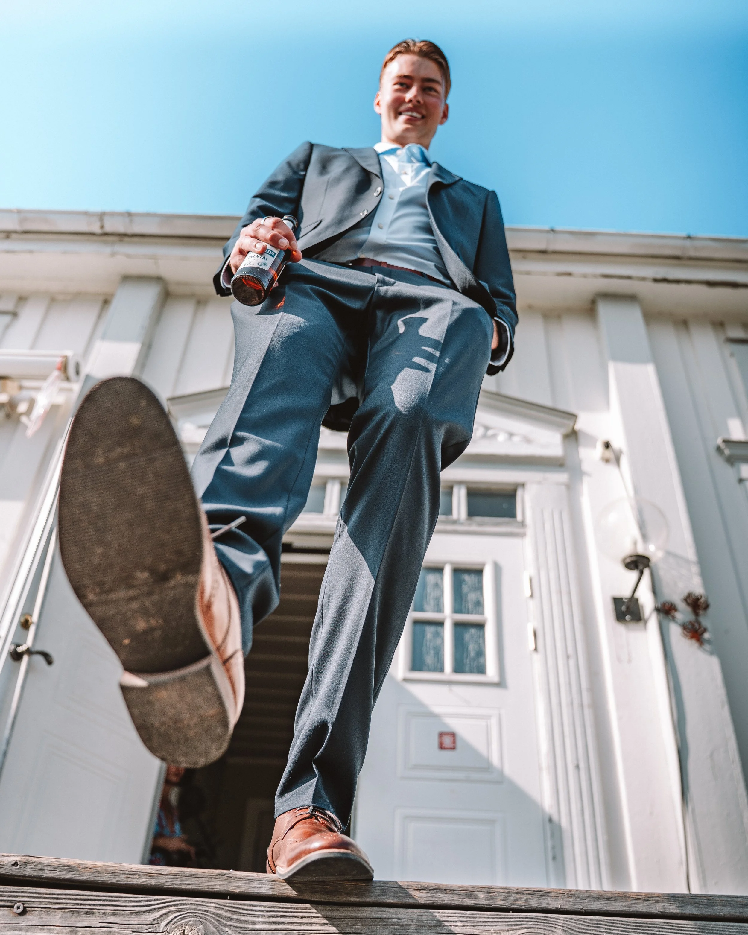 Ung man i kostym som står utanför en vit stuga, tittar ner mot kameran och håller en dryck i handen.