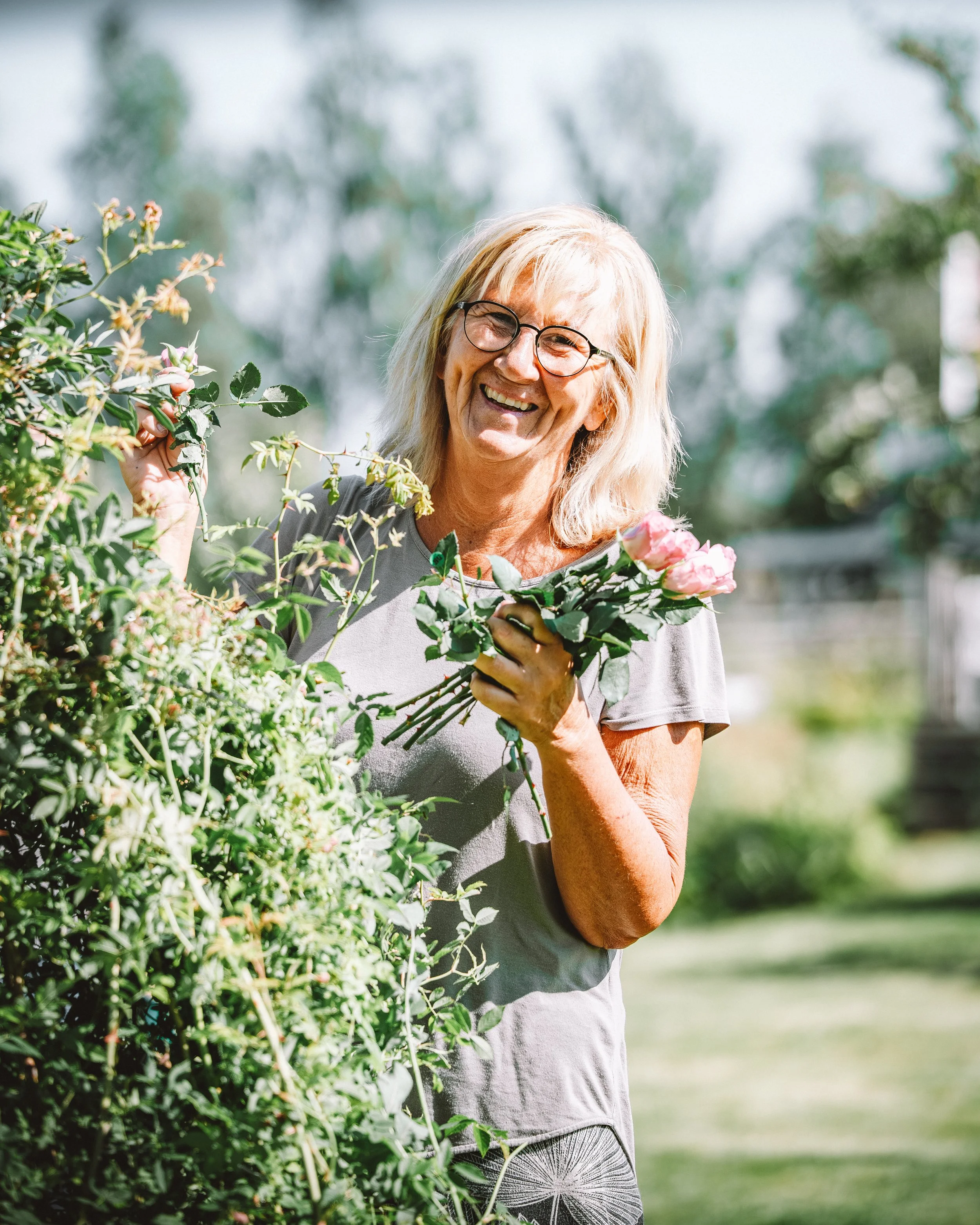 En glad kvinna med glasögon har en bunt rosor i handen, står i en trädgård och skördar blommor.