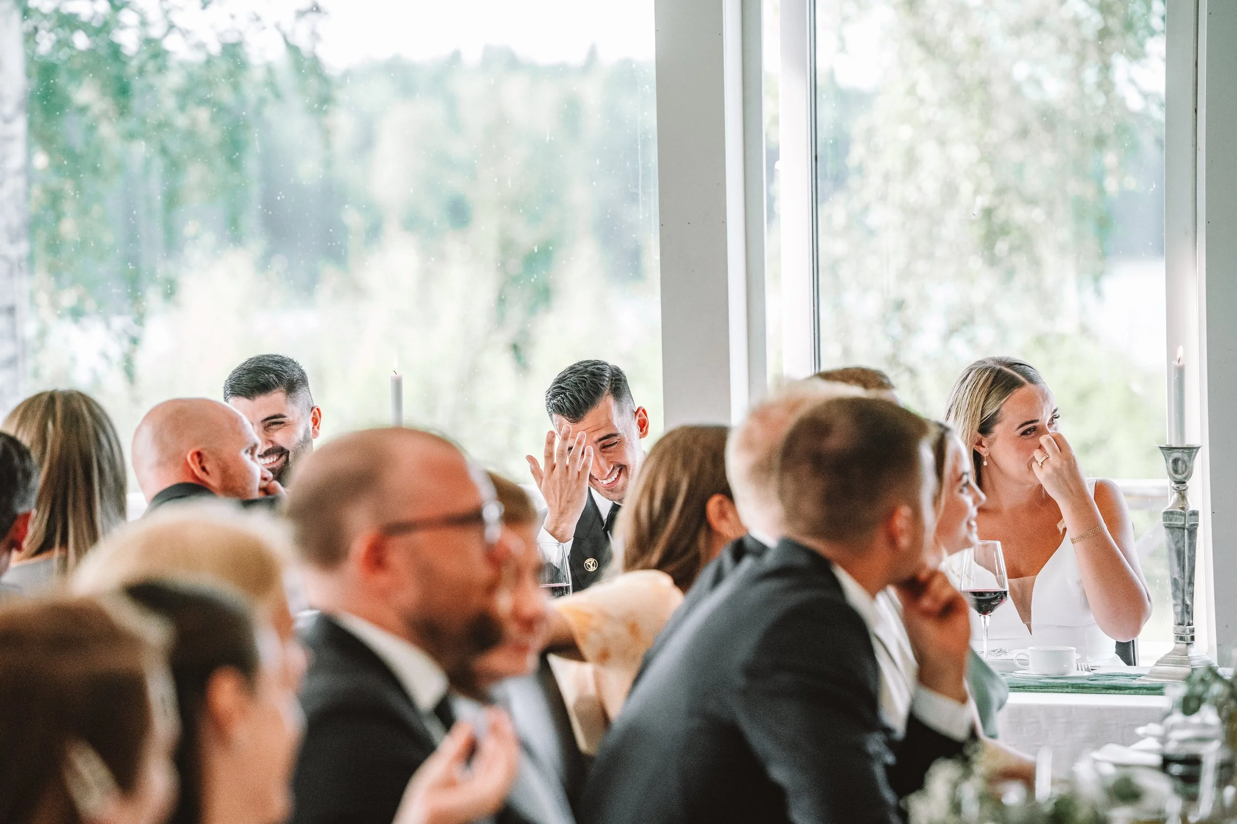 Människor vid festlig tillställning, skratt och samtal, ljus inomhusmiljö, stora fönster med utsikt över grönskog.