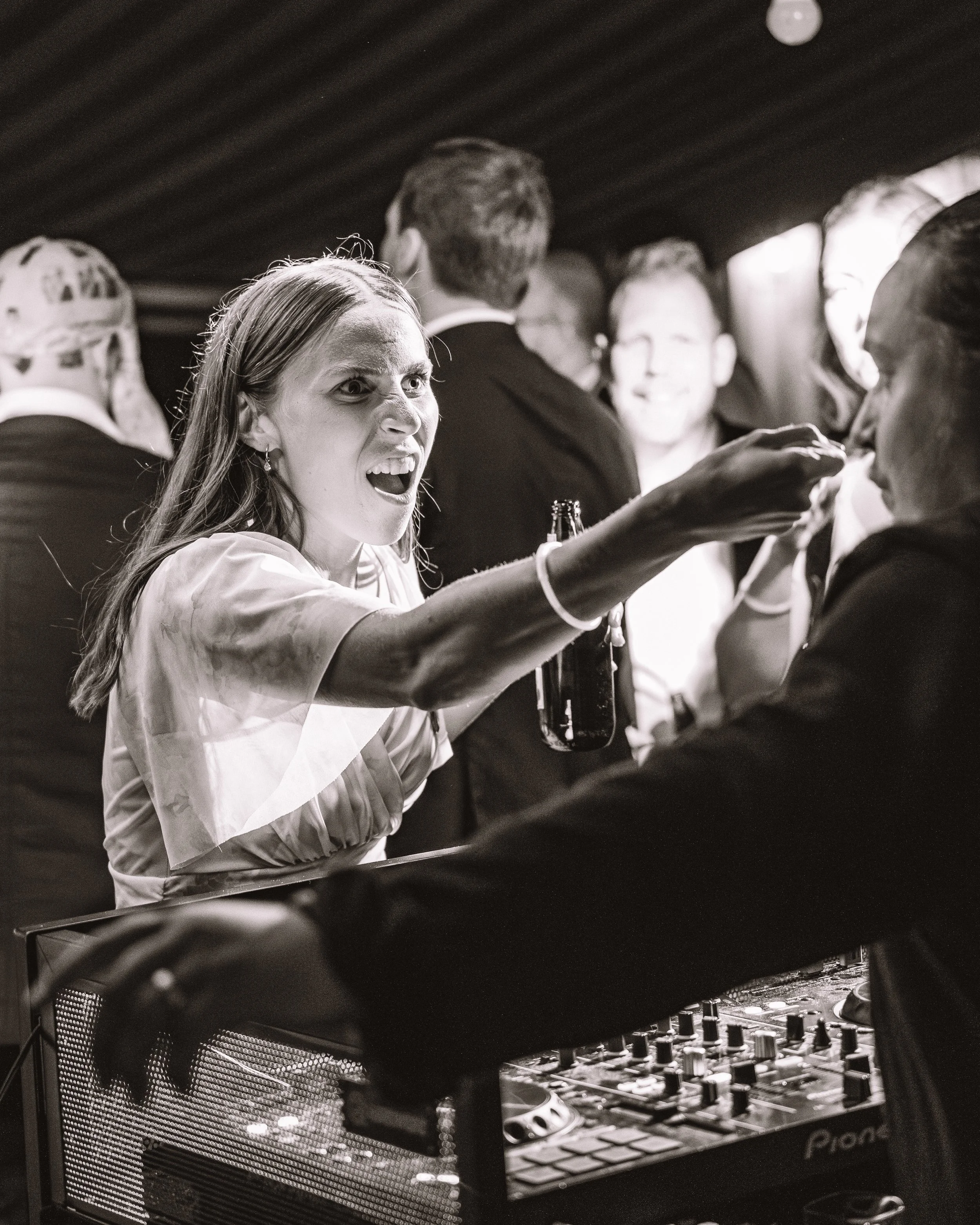 En kvinna med vädjad ansiktsuttryck som pekar med handen mot en person, framför en DJ. Människor i bakgrunden i ett mingel.