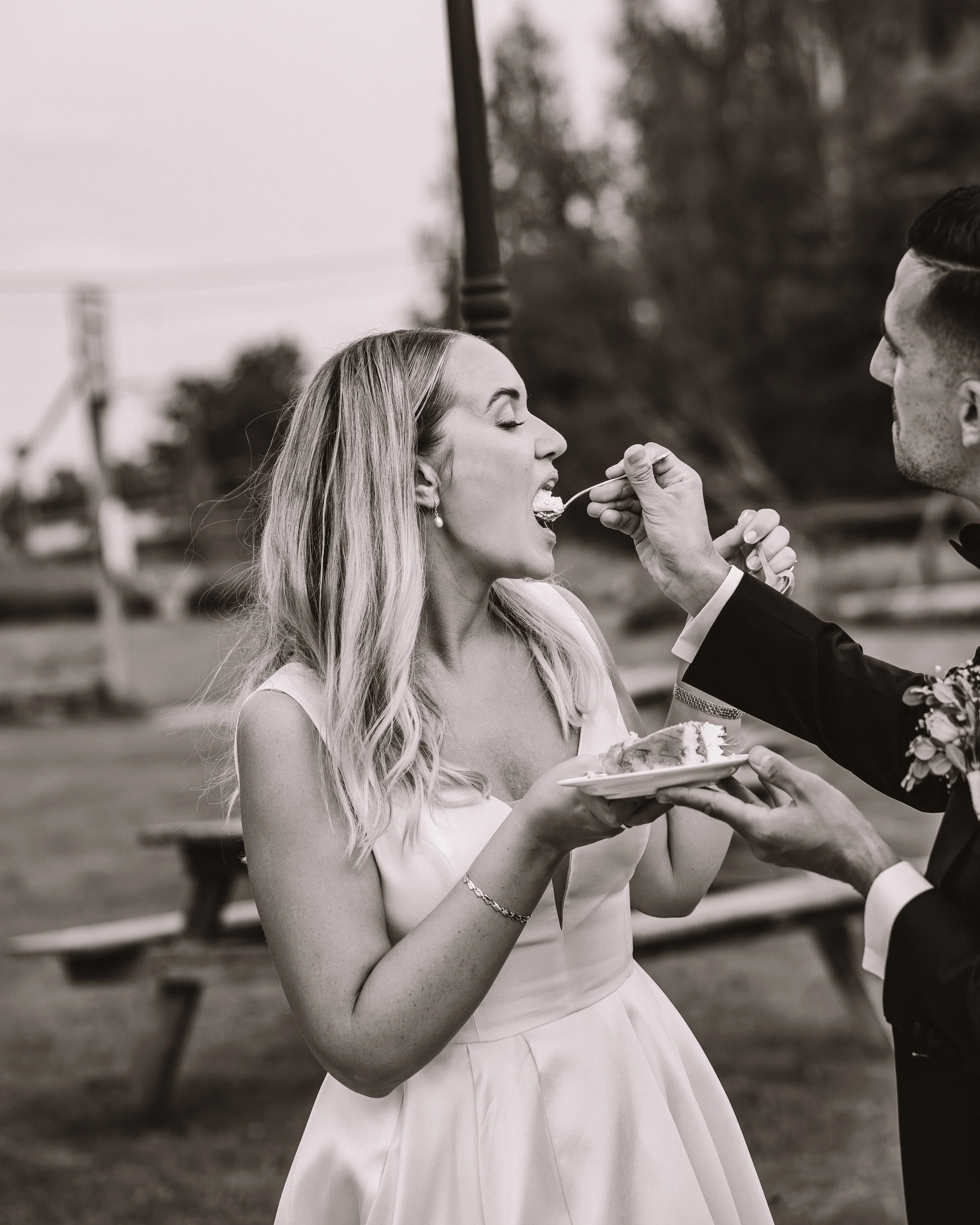 Svartvit bild av ett bröllop där en kvinna i vigselklänning äter tårta som en man i kostym ger henne.