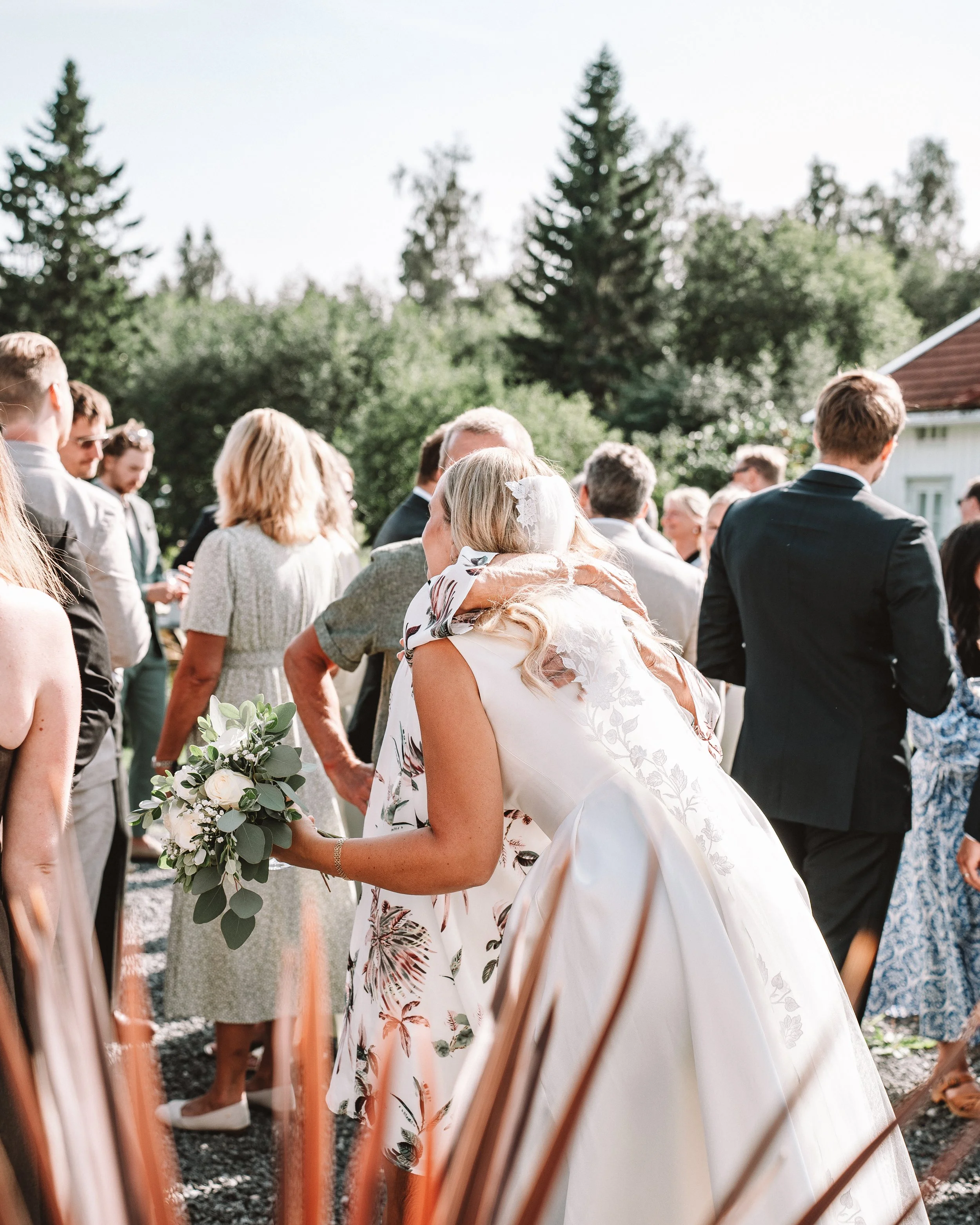En brud med vit klänning och ett flor av gröna blad och vita blommor håller en bukett, omringad av gäster utomhus under en solig dag.