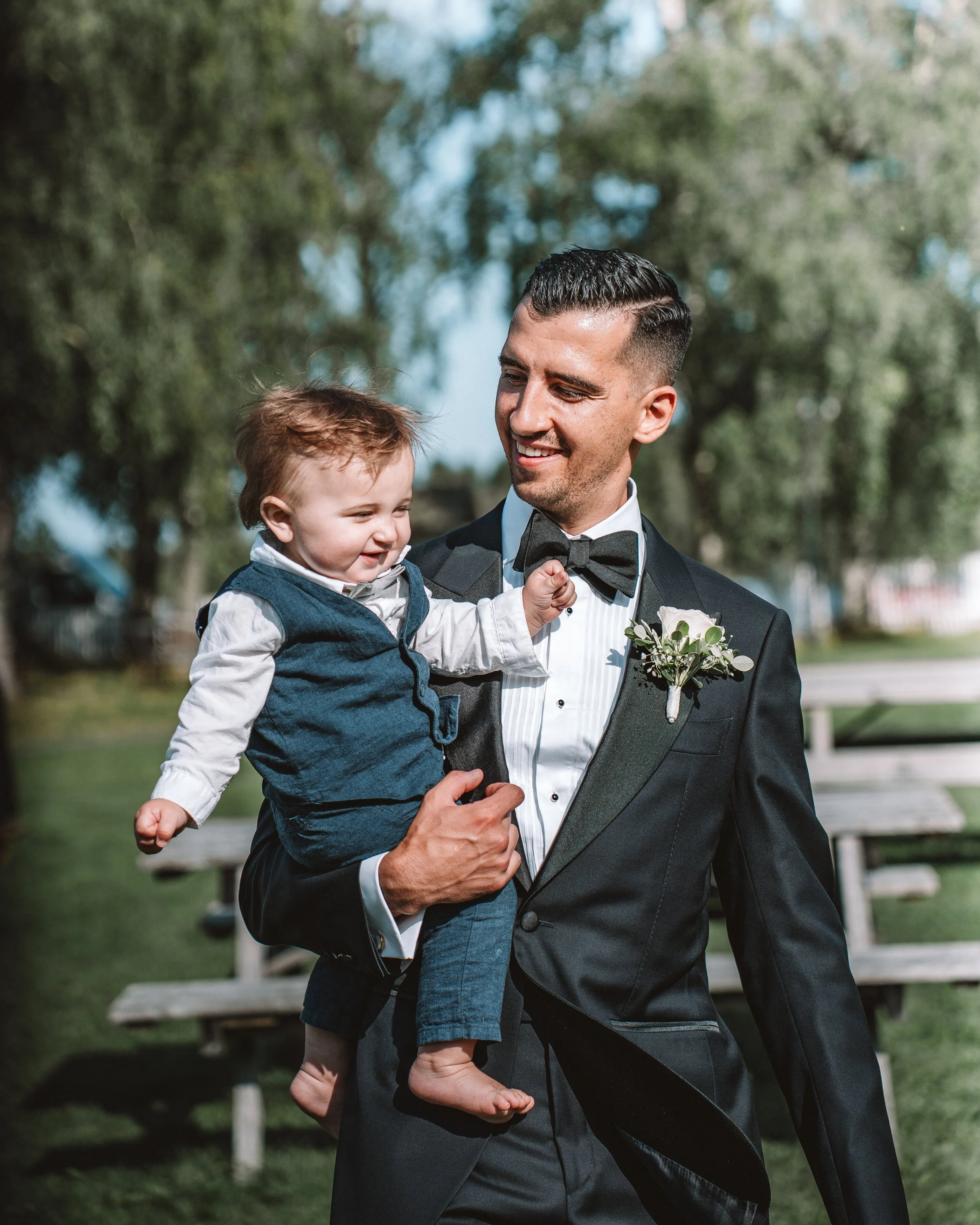 En man i en svart formell kostym bär en liten pojke på axeln, båda ler utomhus.