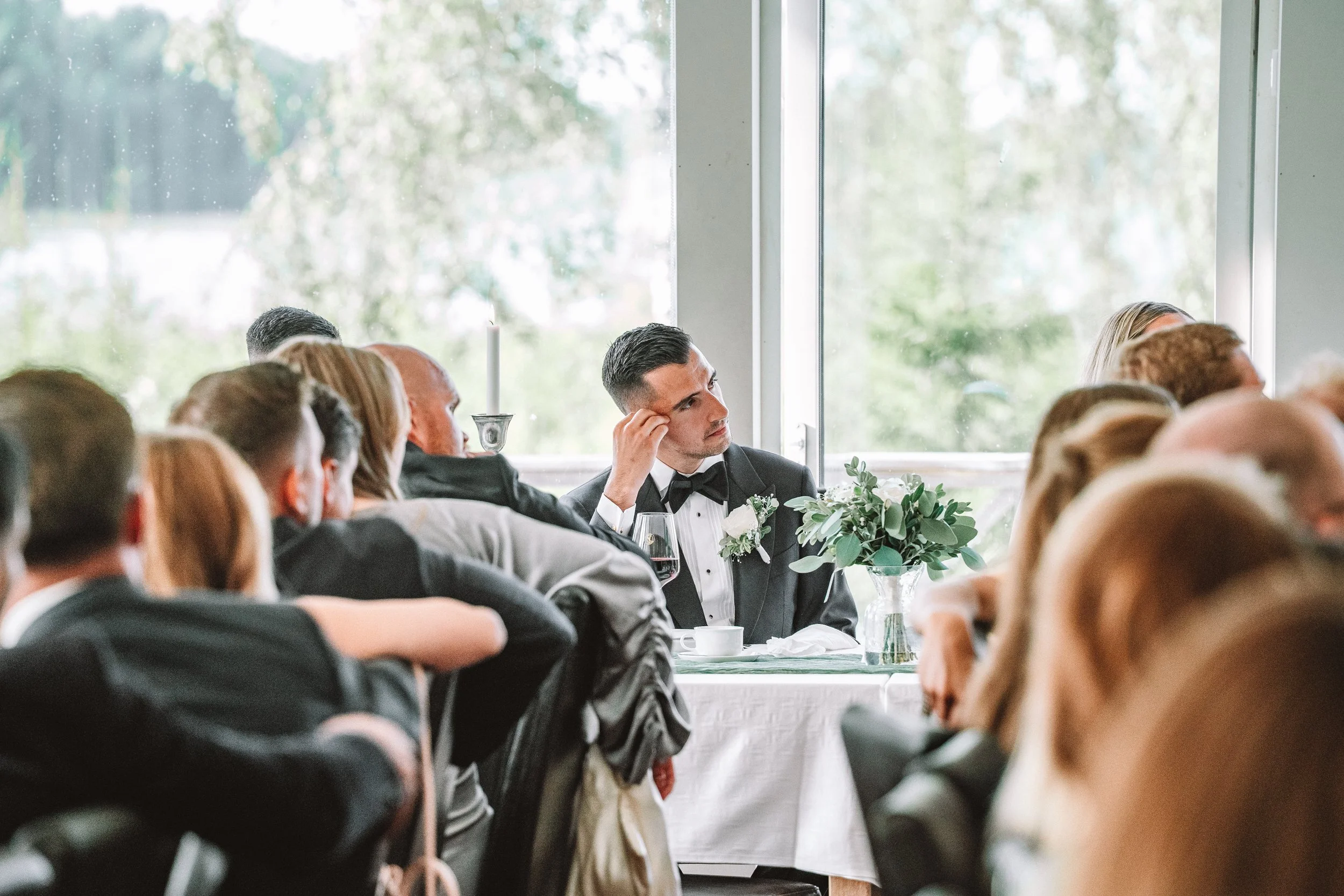 Ännu en grupp vuxna personer i formella kläder, inklusive en man i smoking, som sitter vid ett bord i ett ljusrum med stora fönster, vilket antyder att det är en formell tillställning eller mottagning.