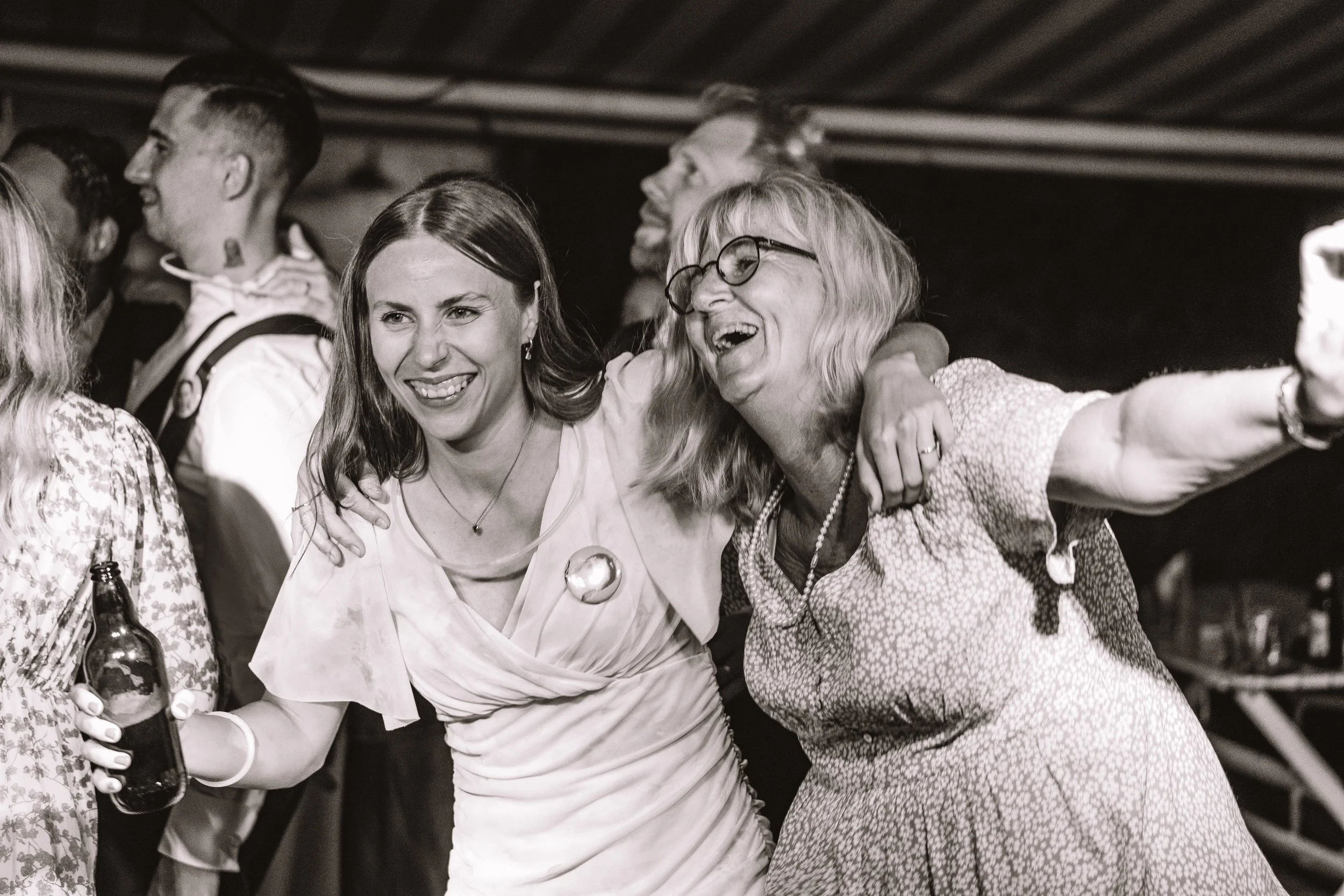 Två kvinnor som ler och skålar, med armar runt varandra i ett socialt sammanhang.