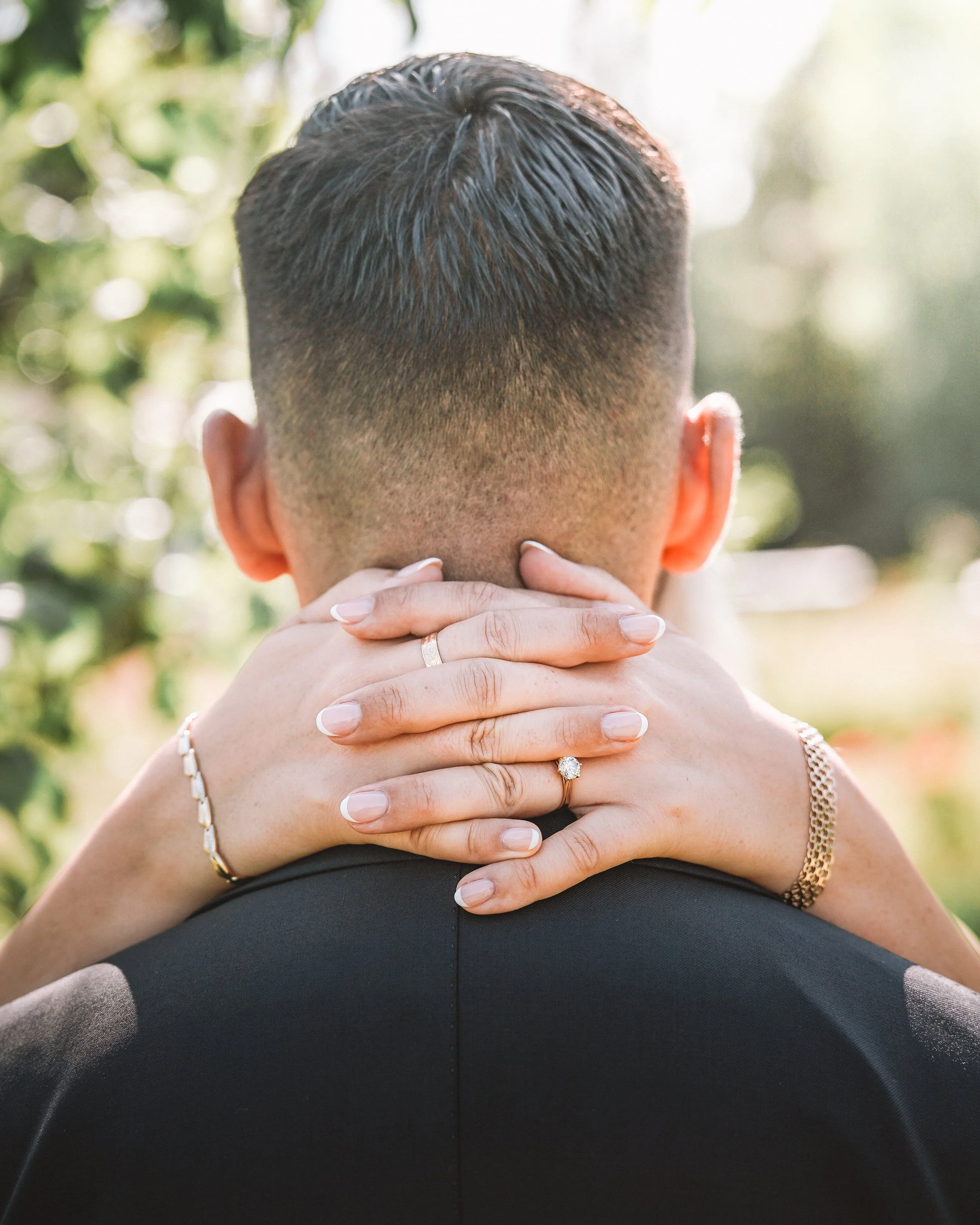 Närbild av en person med kortklippt hår, sett bakifrån, med en kvinna som omfamnar honom från bakom, hennes händer runt hans nacke. Hon bär en vigselring och ett armband.