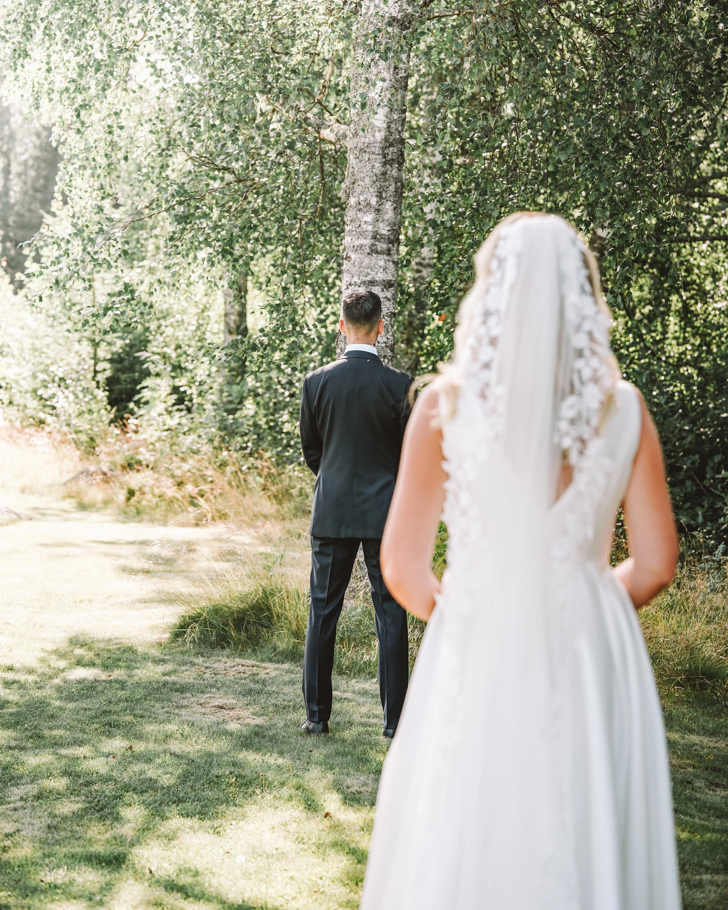 En brud i vitt brudklänning står framför en man i en svart kostym utomhus, omgiven av gröna träd och gräs, i ett skogs- eller parkliknande område.