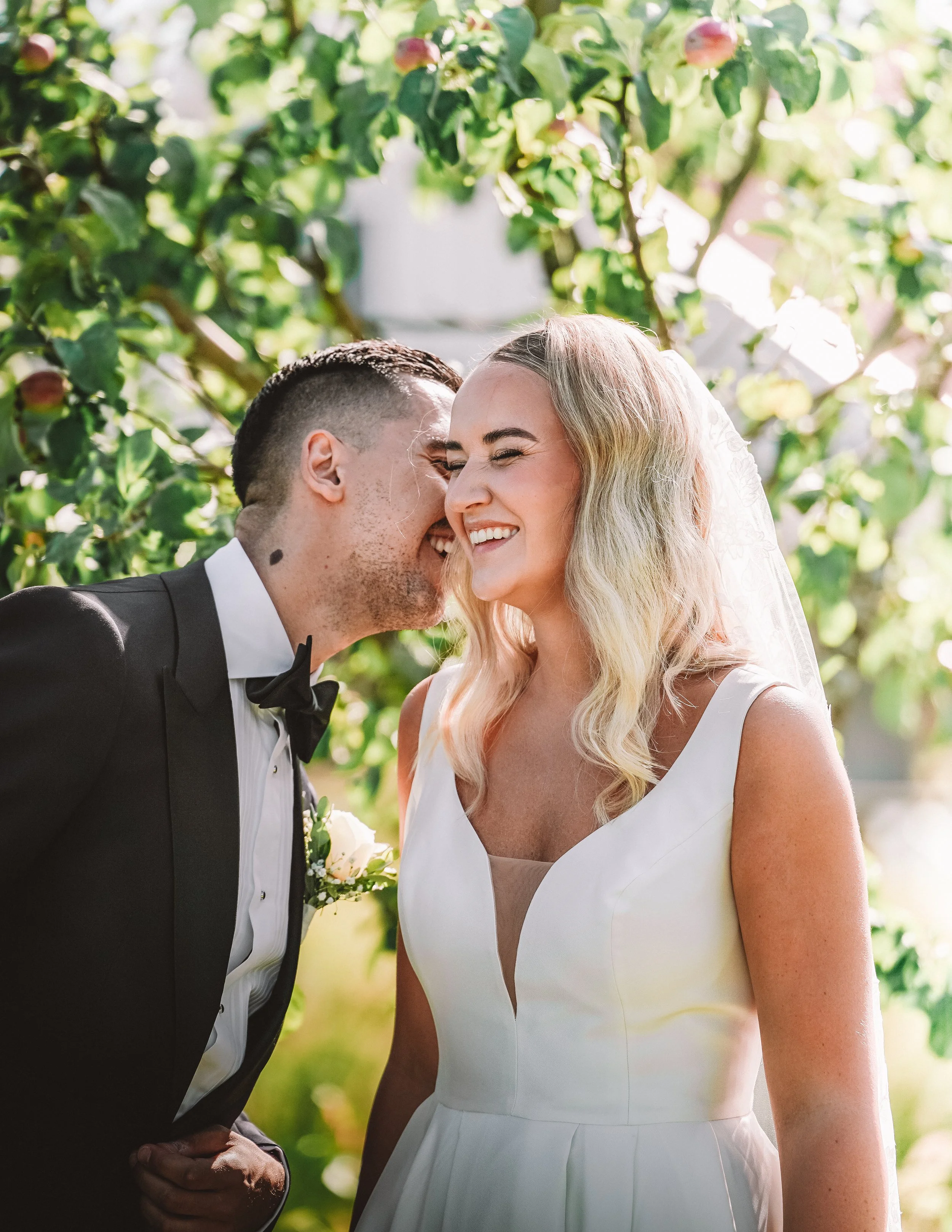 En glad brud och brudgum på bröllop, leende och nära varandra utomhus med gröna löv i bakgrunden.