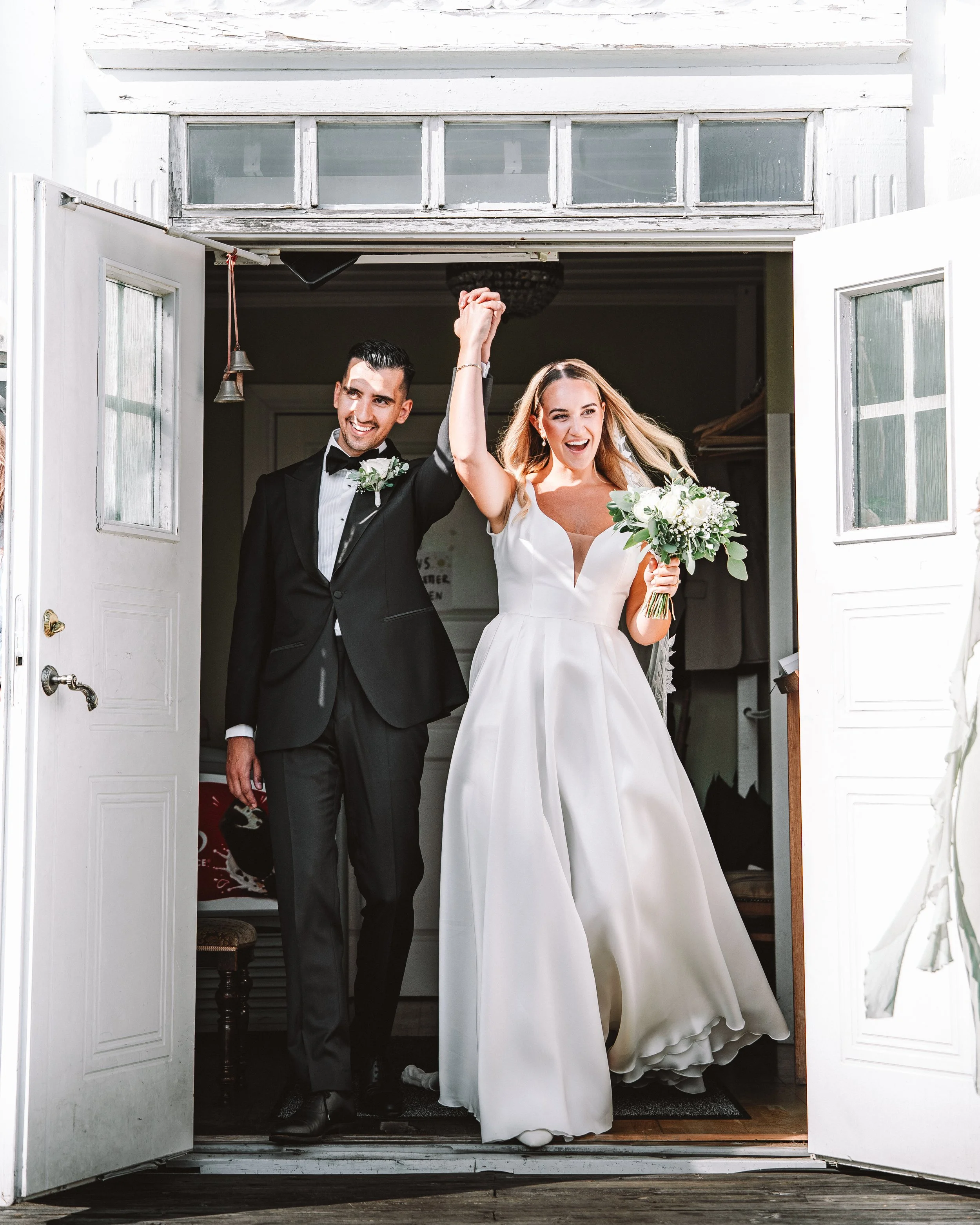 En nygift man och kvinna på bröllopsdag, håller handen uppe i triumf, klädda i formella bröllopskläder, står i ett vitt rum med öppna dörrar.