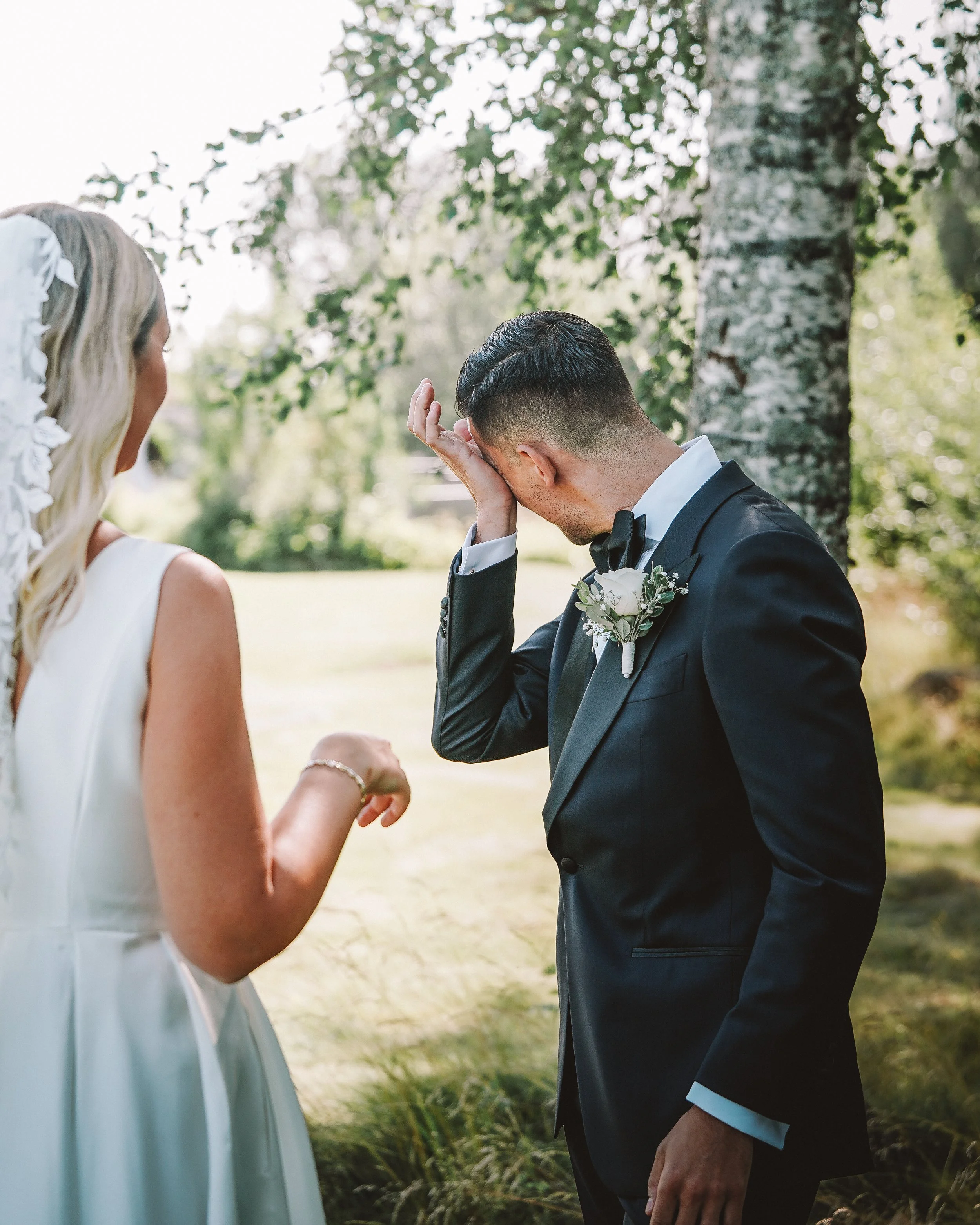 En brud och en brudgum under ett bröllop i en park, där brudgummen torkar sina tårar, med gröna träd och natur i bakgrunden.