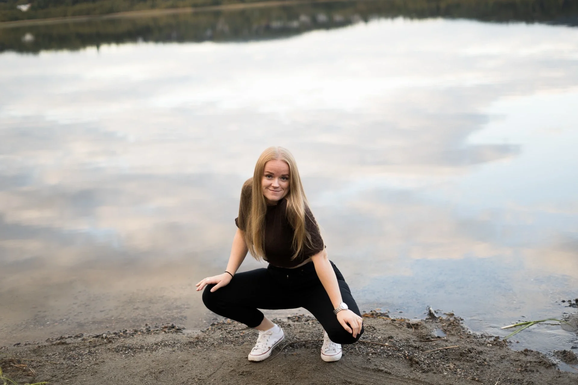 En ung kvinna med långt blont hår, vita sneakers, svart tröja och svarta byxor, som squats vid en sjö, med en lugn vattenyta och en ö i bakgrunden.