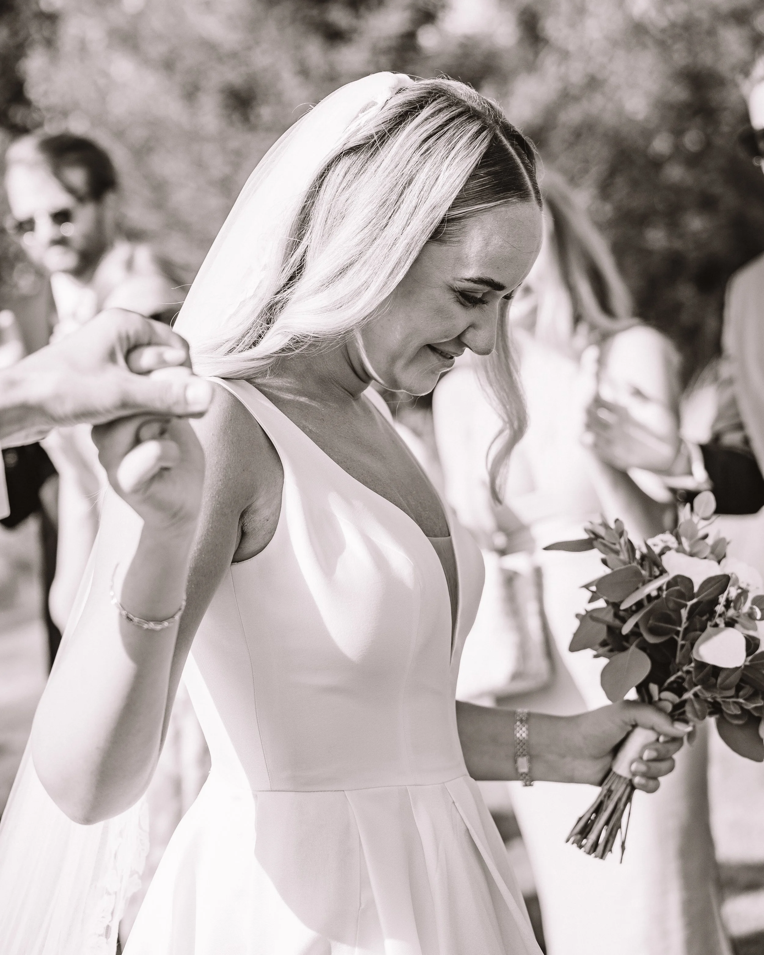 En kvinna i en brudklänning håller en bukett och ler ner medan hon är på en utomhusbröllopsplats.