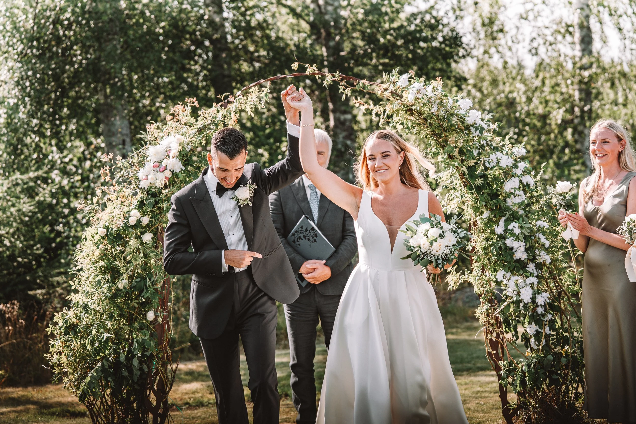 Ett bröllop där en man och en kvinna firar äktenskap utomhus, kvinna i vit brudklänning och man i svart tuxedo, under en blomsterdekoration, med gäster runtom.