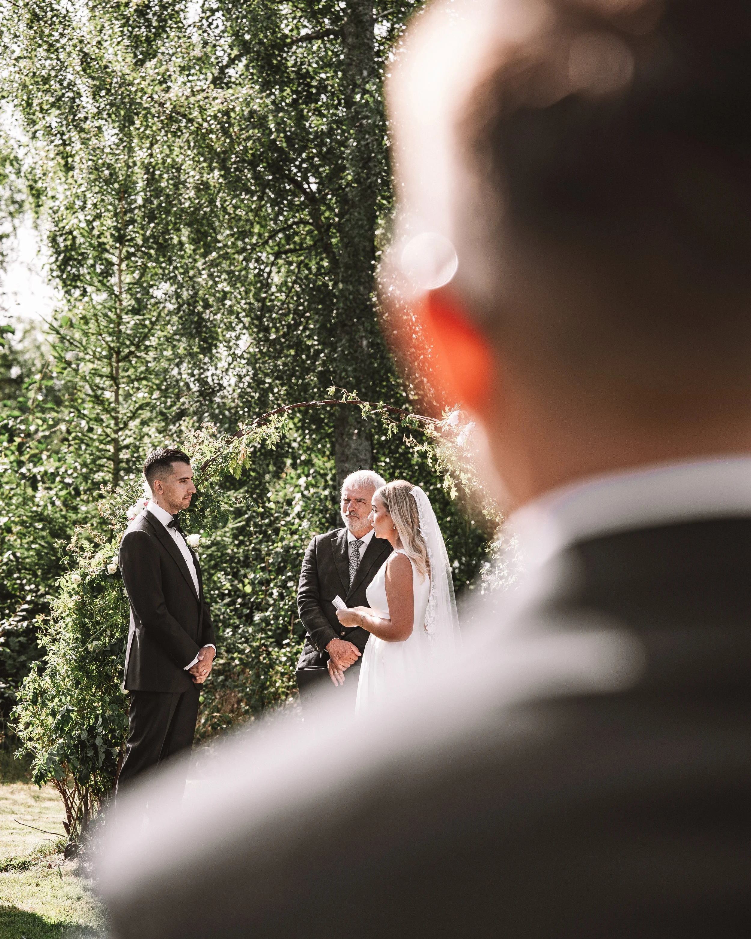 En bröllopsceremoni utomhus med tre personer i fokus, en brud i vit klänning och slöja, en groom i svart kostym, och en officiant i svart kostym, omgivet av gröna träd och solsken, med en annan person vars ansikte är delvis synligt i förgrunden.