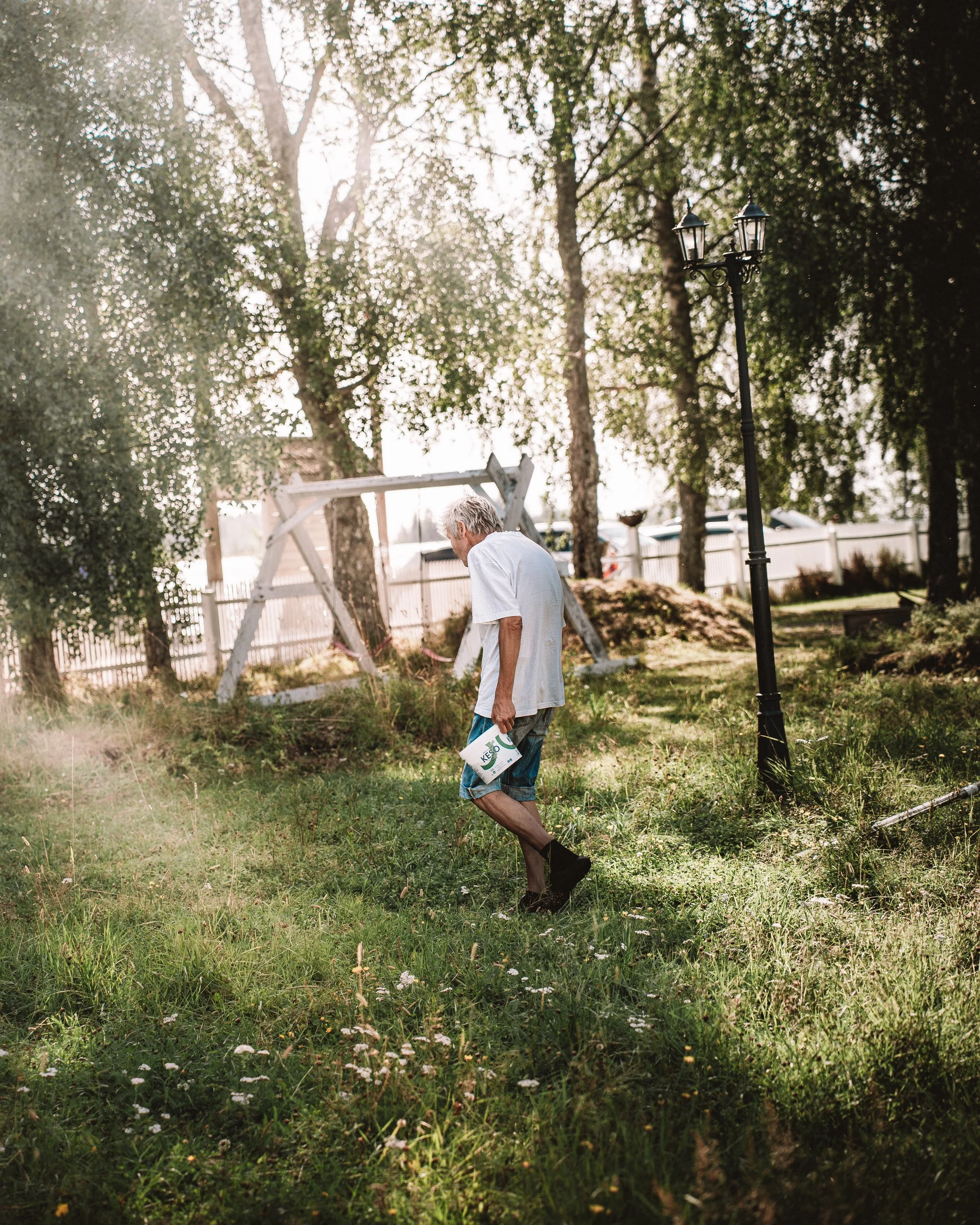 En äldre man i vita tröja och shorts går på en gräsmatta med en bricka i handen i ett trädgårdsområde med träd och en lekplats