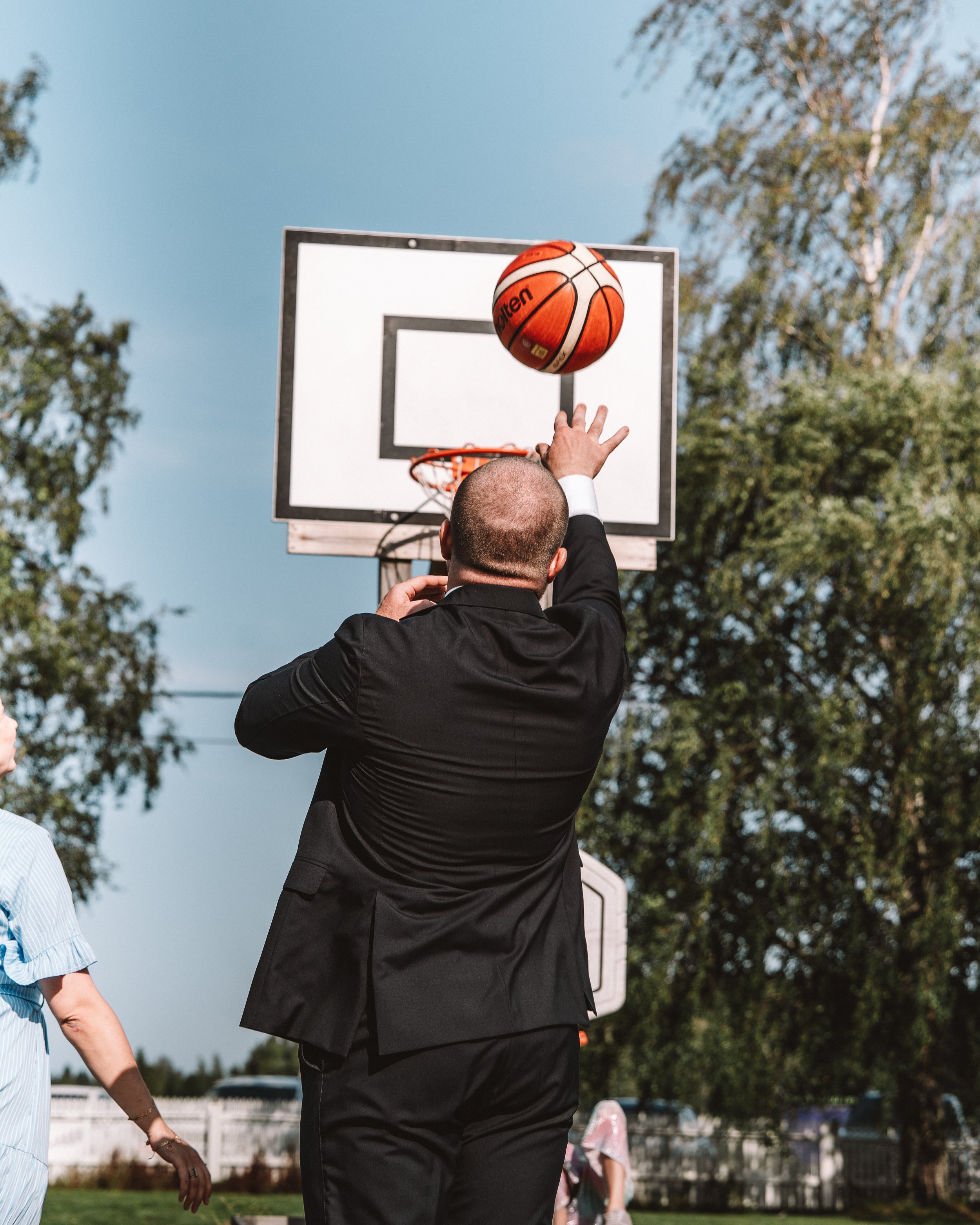 En man i svart kostym skjuter en basketboll mot korgen utomhus.