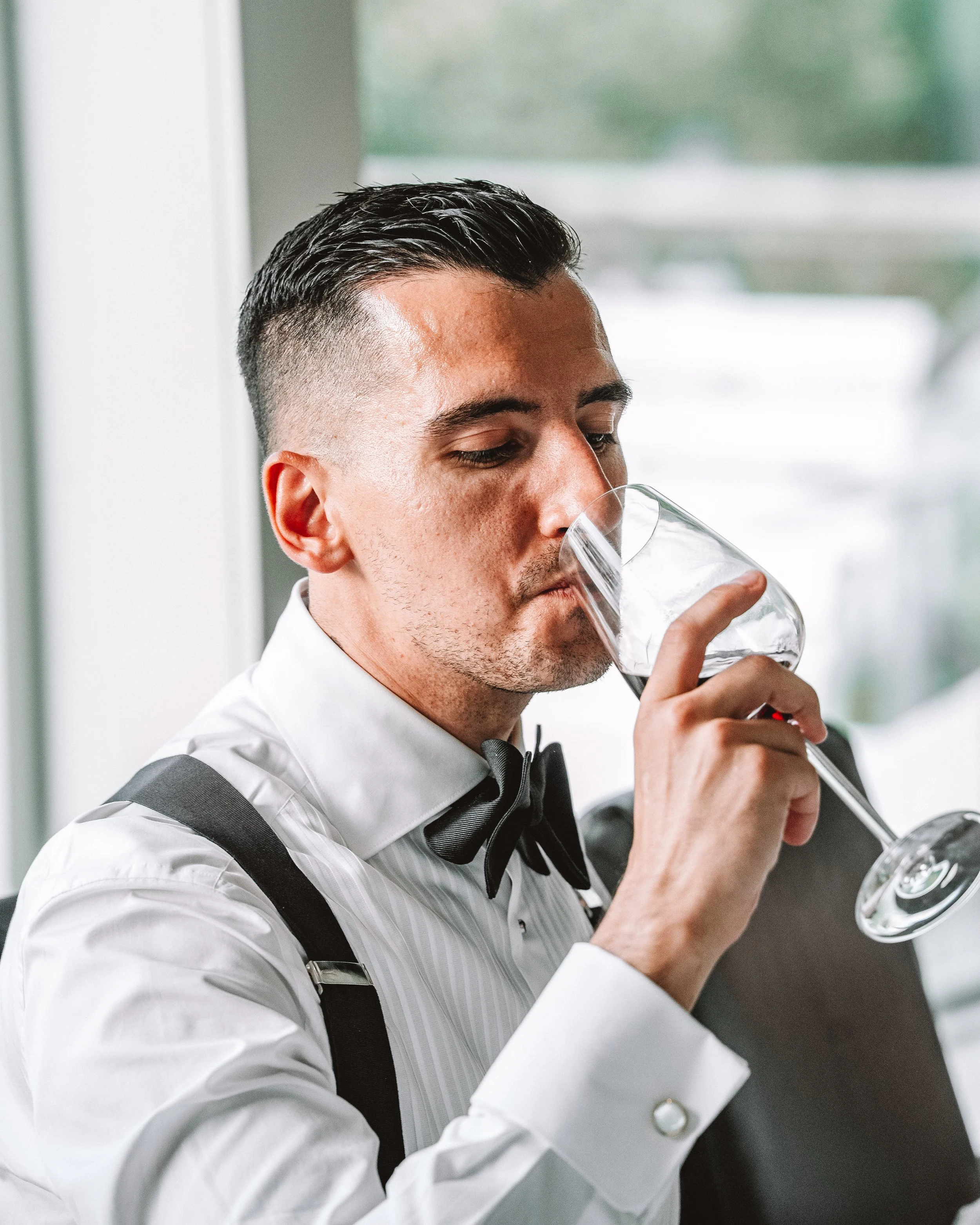 En man i vit skjorta, svart fluga och mörka hängslen dricker vin ur ett vinglas.