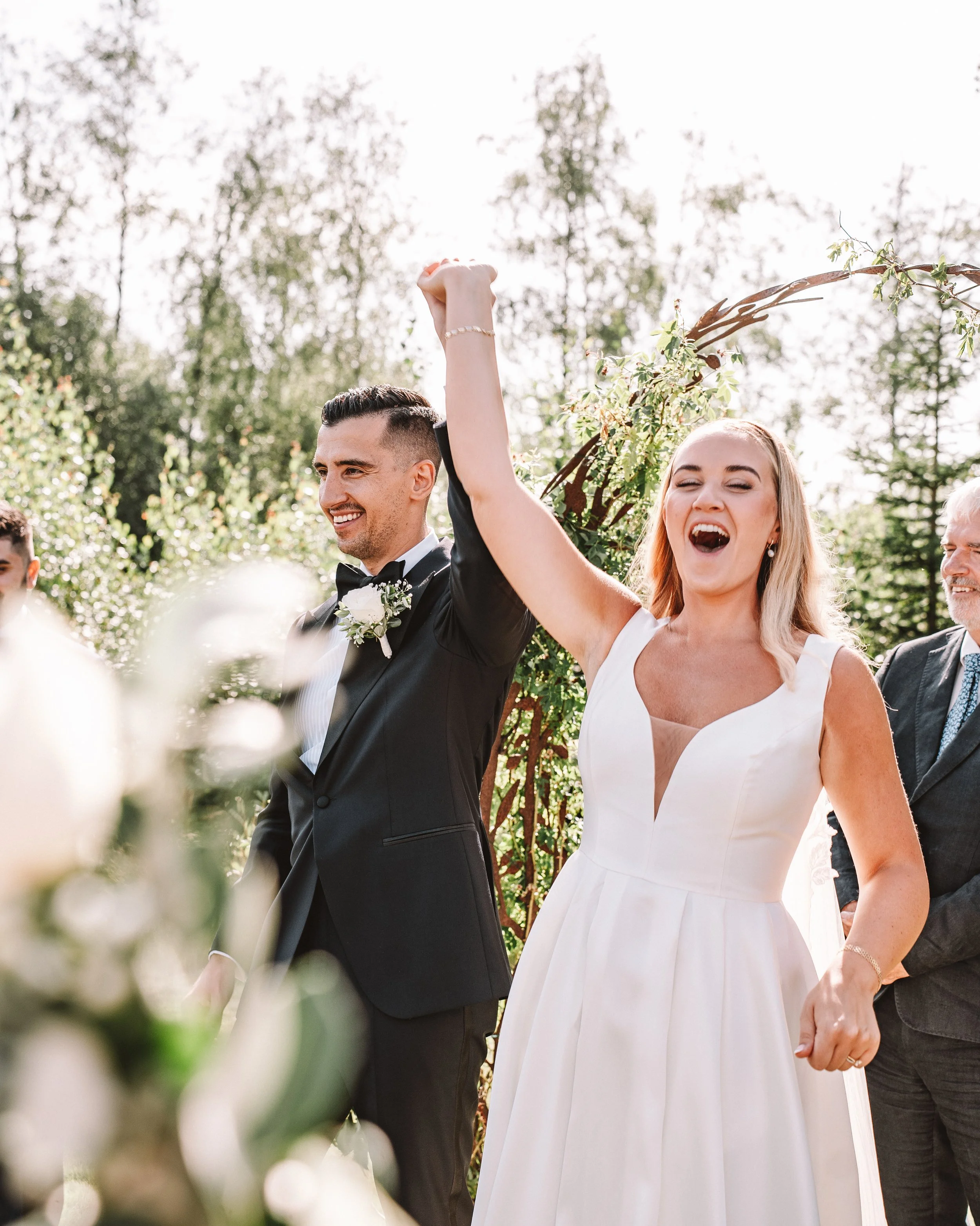 Brudpar firar bröllop utomhus, kvinna i vit brudklänning och man i svart kostym med slöja, viftar med handen, glada ansikten.