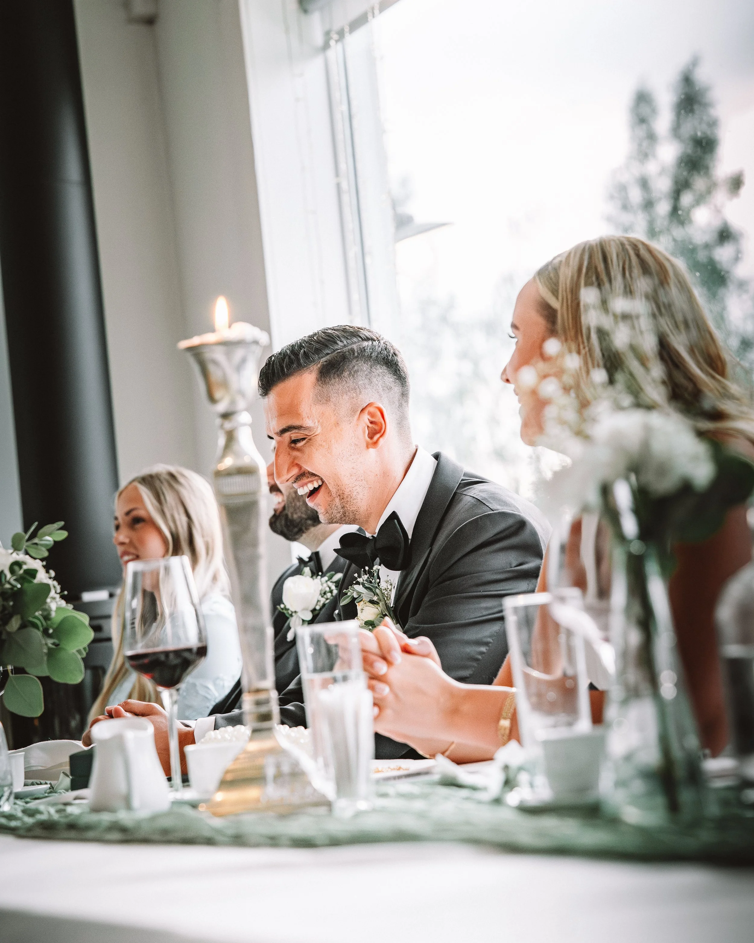 Män och kvinnor vid bord under ett bröllop, smiling, med blommor och ljus, nära ett fönster.