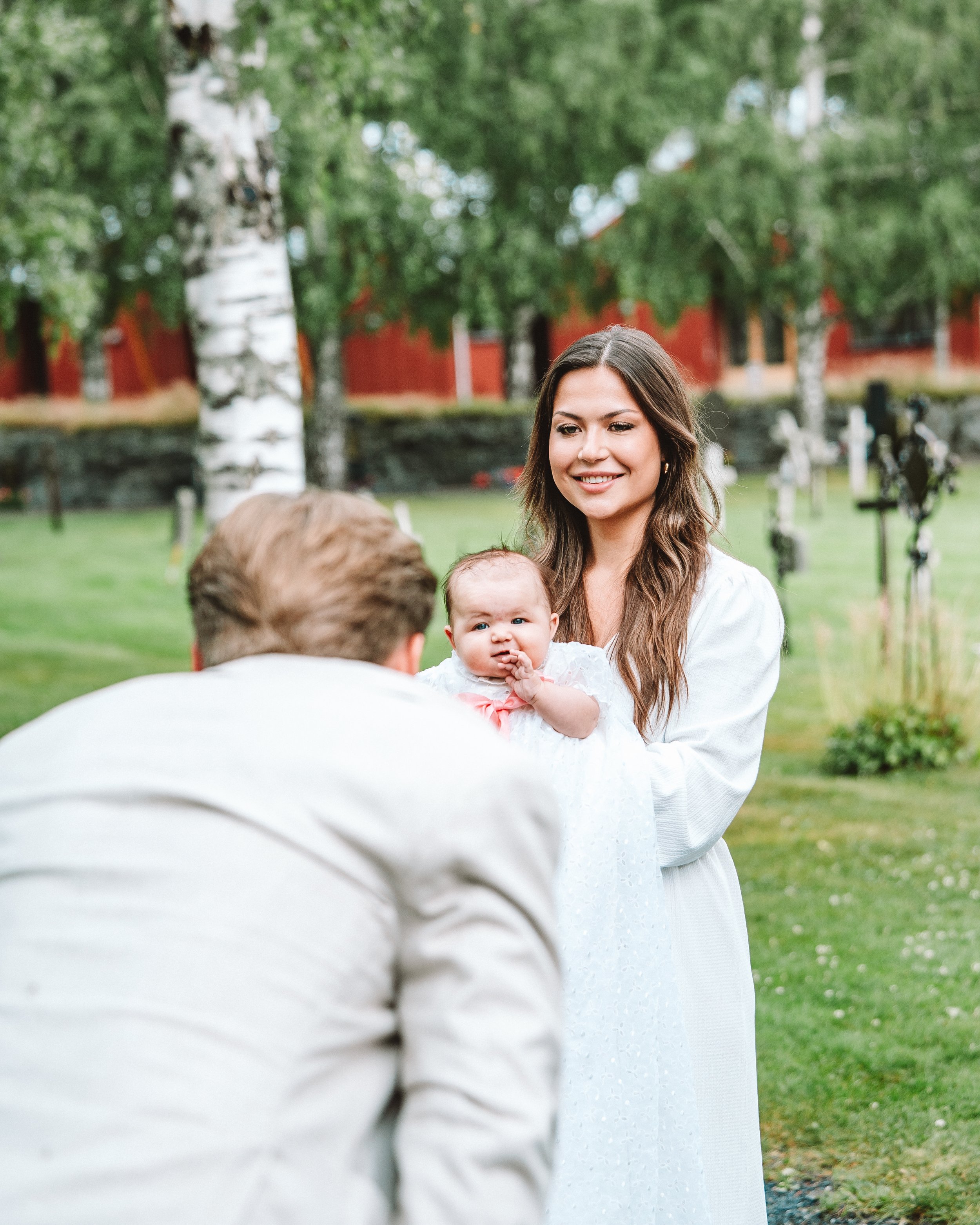 En kvinna håller ett barn i en trädgård efter ett dop i Örnsköldsvik, Höga Kusten, mannen böjer sig ner framför dem och grimaserar mot barnet, bakgrund med gröna träd och en röd byggnad.