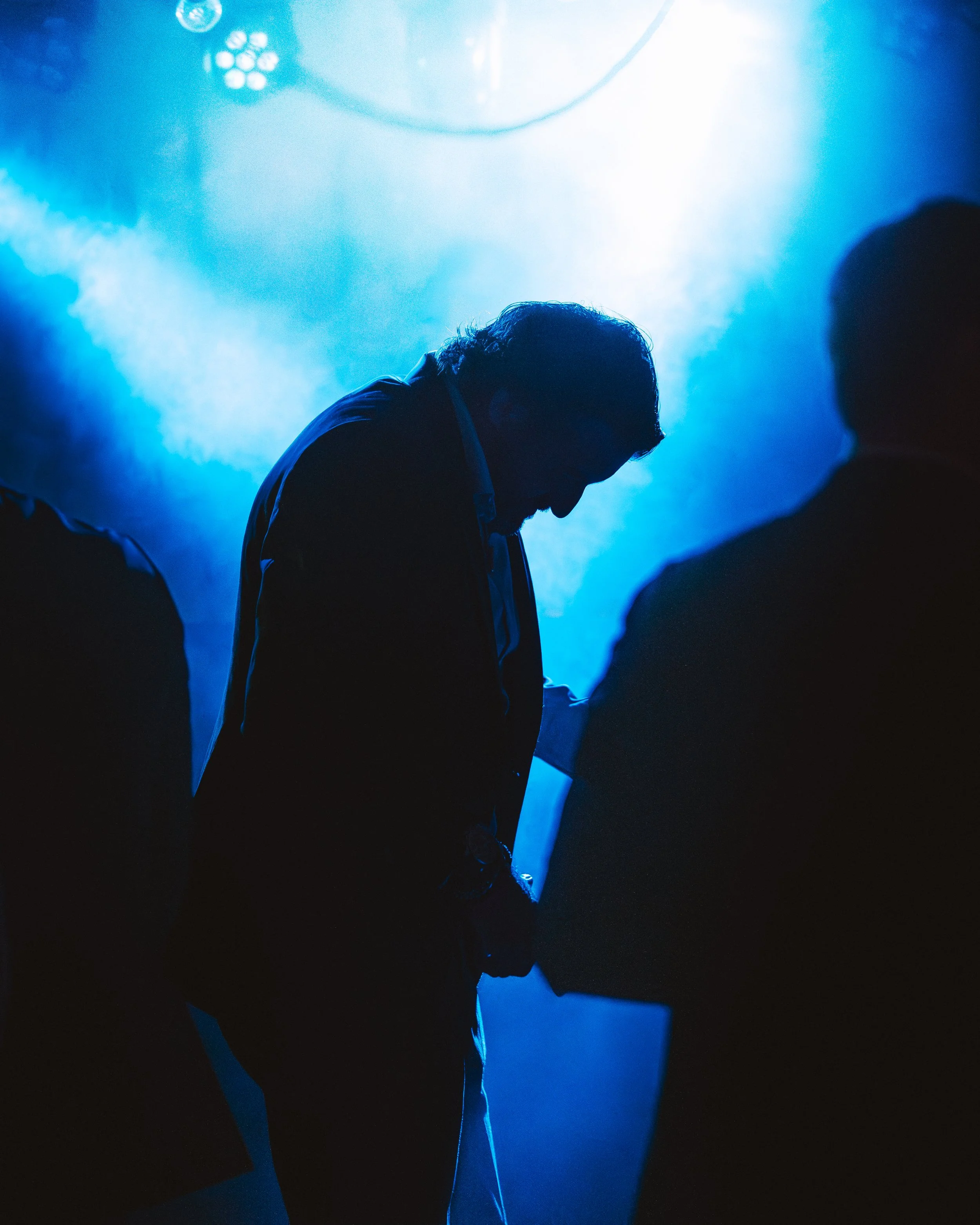 Silhuetter av tre personer i blå belysning, en person i fokus ser nedåt med huvudet lutat, bakgrund av rök och ljus.